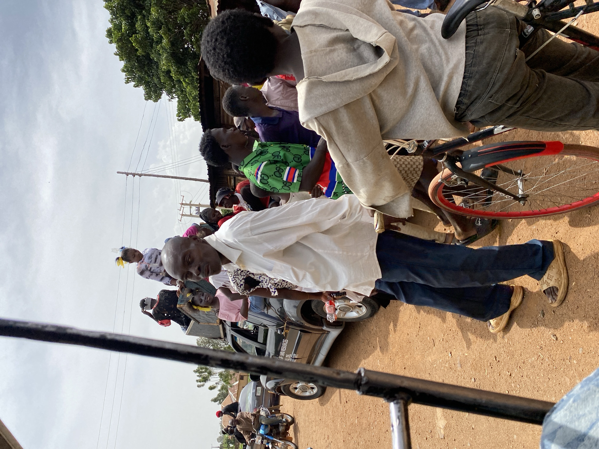 A daytime scene in Kuru Dungus, Nigeria shows a gathering of people in an outdoor environment. A dark gray car with lettering "RESOURCES" partially visible on its side is present. People are standing around the car and in the background. A man wearing a white shirt and blue pants stands prominently in the foreground. Another individual on a bicycle with a red rim is also visible. The ground is reddish-brown. A tree is visible in the background. Power lines stretch across the sky.