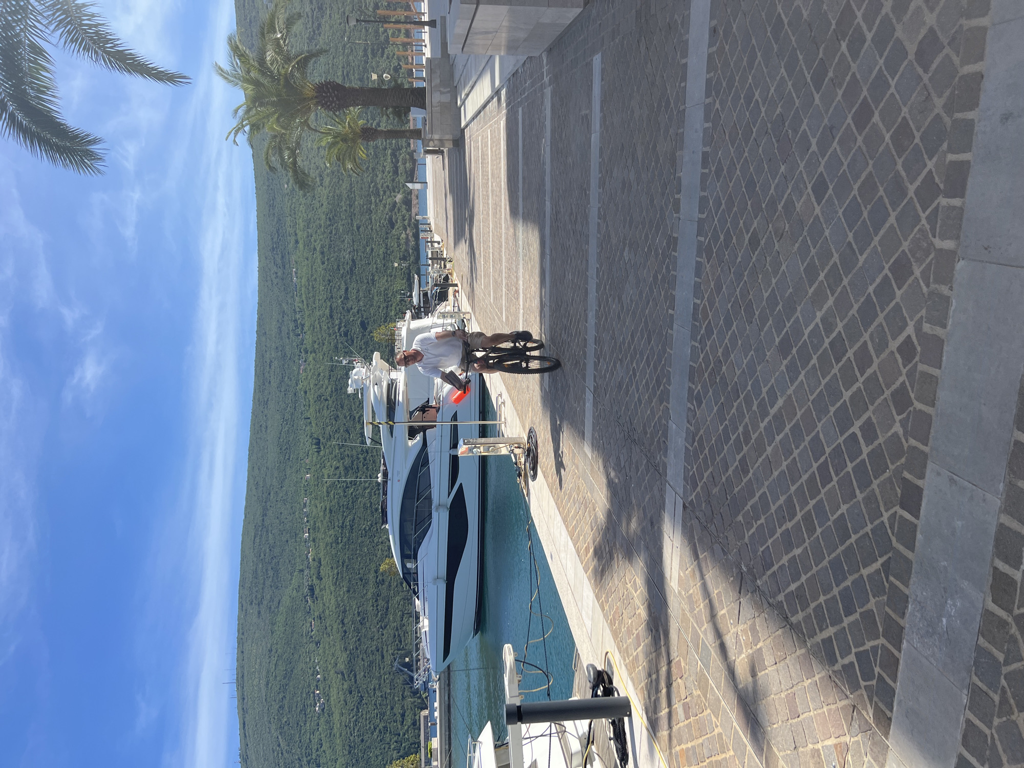 The image shows a sunny day on a paved waterfront promenade in Herceg Novi, Montenegro. A man is riding a bicycle along the promenade. A large white yacht is docked along the waterfront. In the background, a tree-covered mountain range meets a blue sky with light clouds. Palm trees are visible near the promenade.