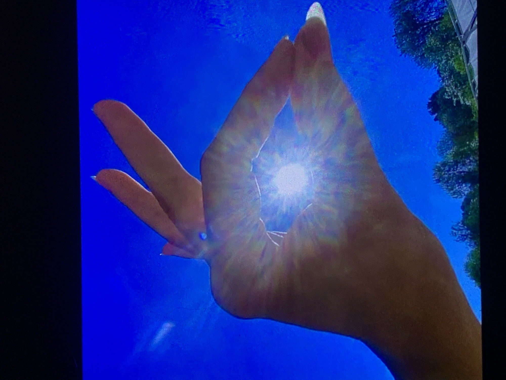 A person's hand is held up against a bright blue sky, with two fingers and a thumb forming a circle. The sun is shining through the opening, creating a lens flare effect with visible rays. The background shows a green landscape with trees and a rooftop below. The image is a close-up shot, with the top portion of the frame obscured by a black area, possibly a television screen. There are no discernible details to indicate the location in Sevastopol, Ukraine.
