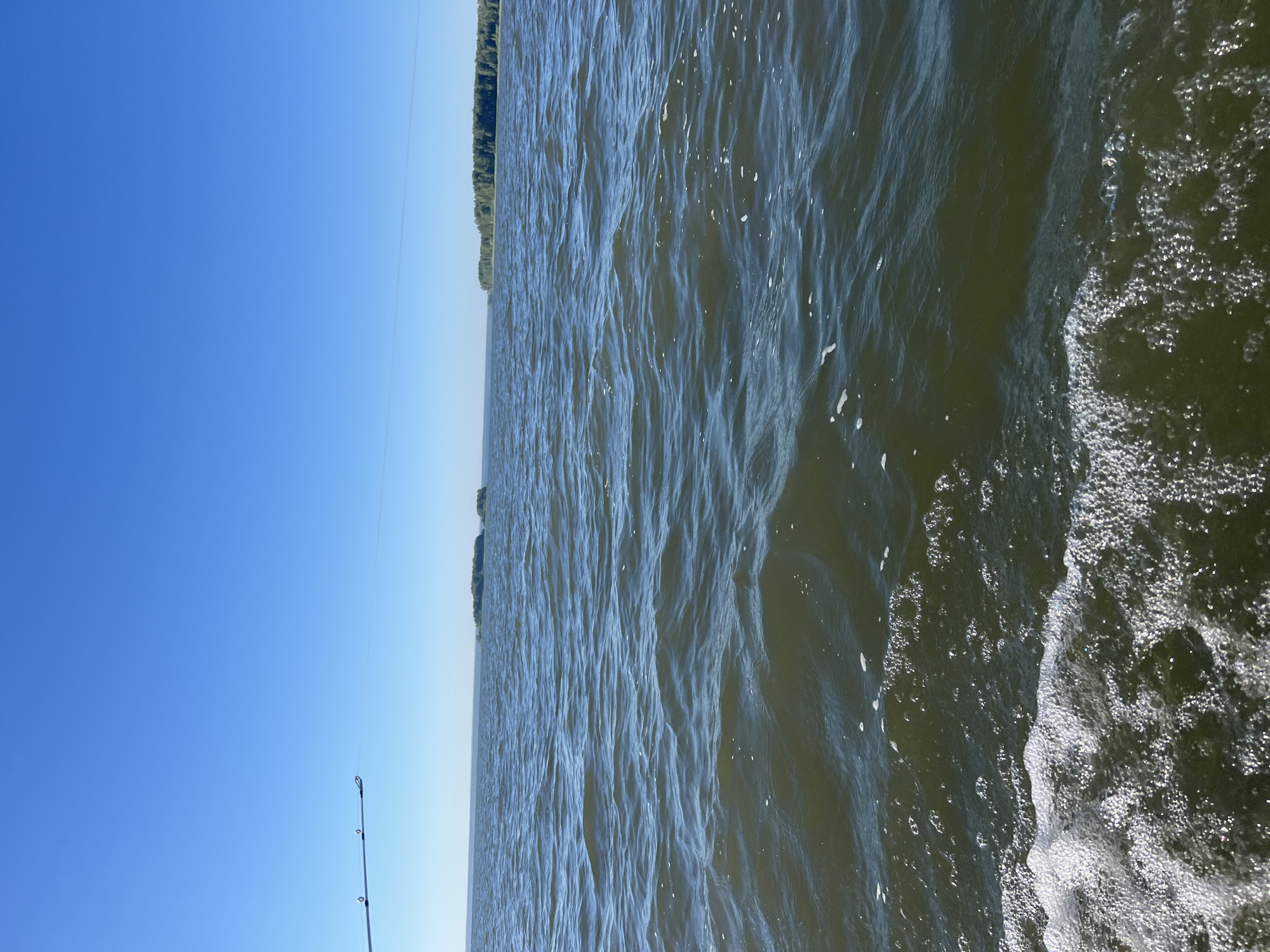 A wide-angle shot captures a body of water under a clear blue sky. In the foreground, a fishing rod extends from the left side of the frame, its tip and line visible against the sky. The water is a dark, greenish-brown, with small waves creating ripples and some white foam near the bottom edge of the image, indicating movement, likely from a boat. In the distance, two small, tree-covered islands are visible, and further still, a long stretch of coastline covered in dense forest. The horizon is sharp and clear, suggesting good visibility. The overall scene evokes a sense of calm and outdoor activity. The location is the confluence of the Yenisei River and the Kan River in Novopichugovo, Russia.