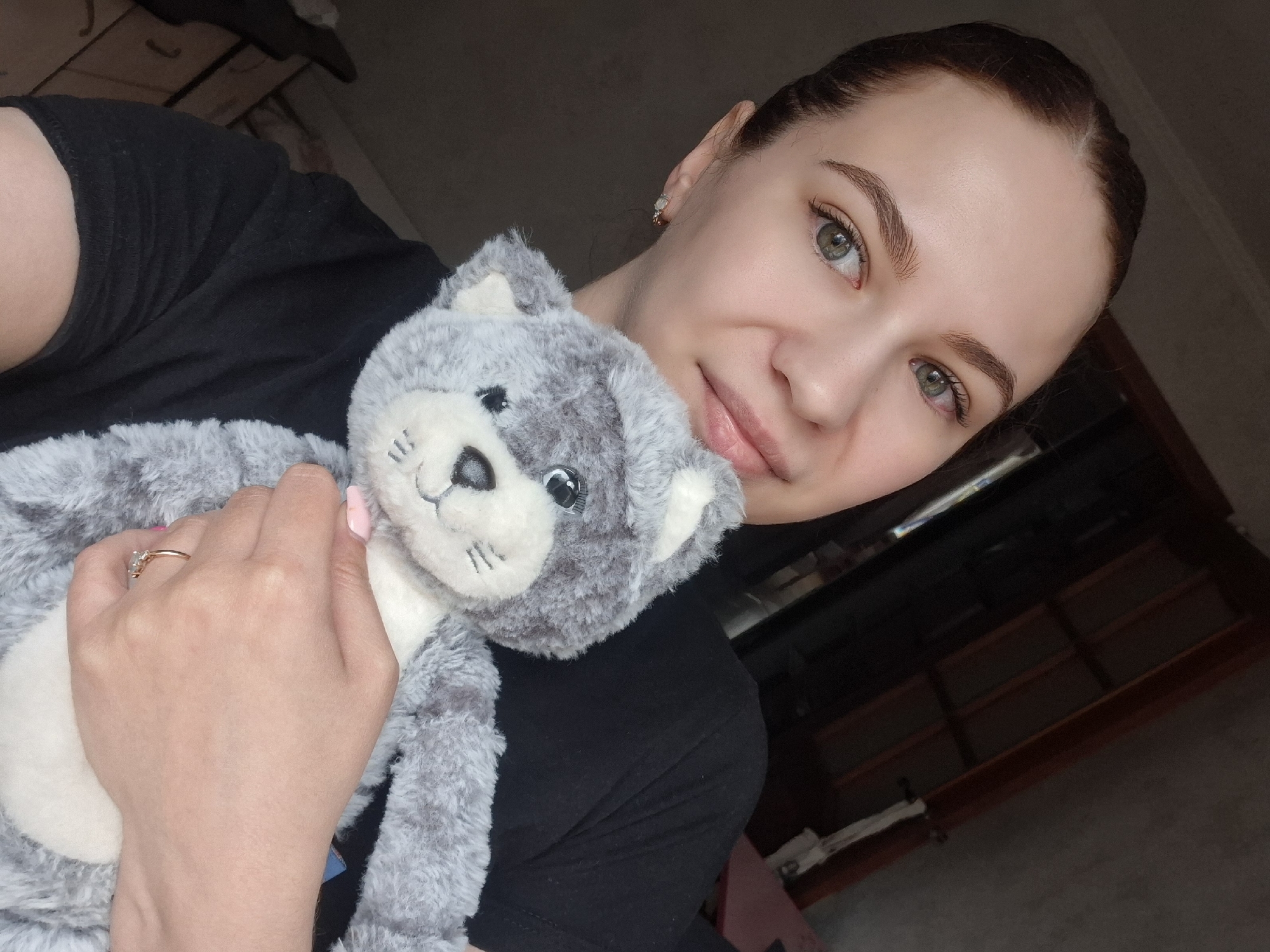 A young woman is captured in a close-up, slightly tilted portrait, holding a plush toy. She has fair skin, prominent green eyes, and dark hair pulled back from her face, revealing her forehead and ears. She wears a black t-shirt and small, light-colored stud earrings are visible in her left ear. Her lips have a natural, glossy appearance, and she offers a subtle, gentle smile towards the viewer.
She is holding a soft, gray and white stuffed animal, resembling a cat or a wolf cub, with large black eyes and embroidered facial features. Her left hand, adorned with a ring featuring multiple small stones on her ring finger, is gently cradling the toy. Her fingernails are painted a light pink.
The setting appears to be indoors, possibly a room. The background is dimly lit and largely out of focus, showing dark, indistinct shapes that suggest furniture or shelving behind her. The overall lighting suggests natural light coming from an unseen source, illuminating her face and the toy. No text is visible in the image. The context for the location is Chaikovsky, Russia.