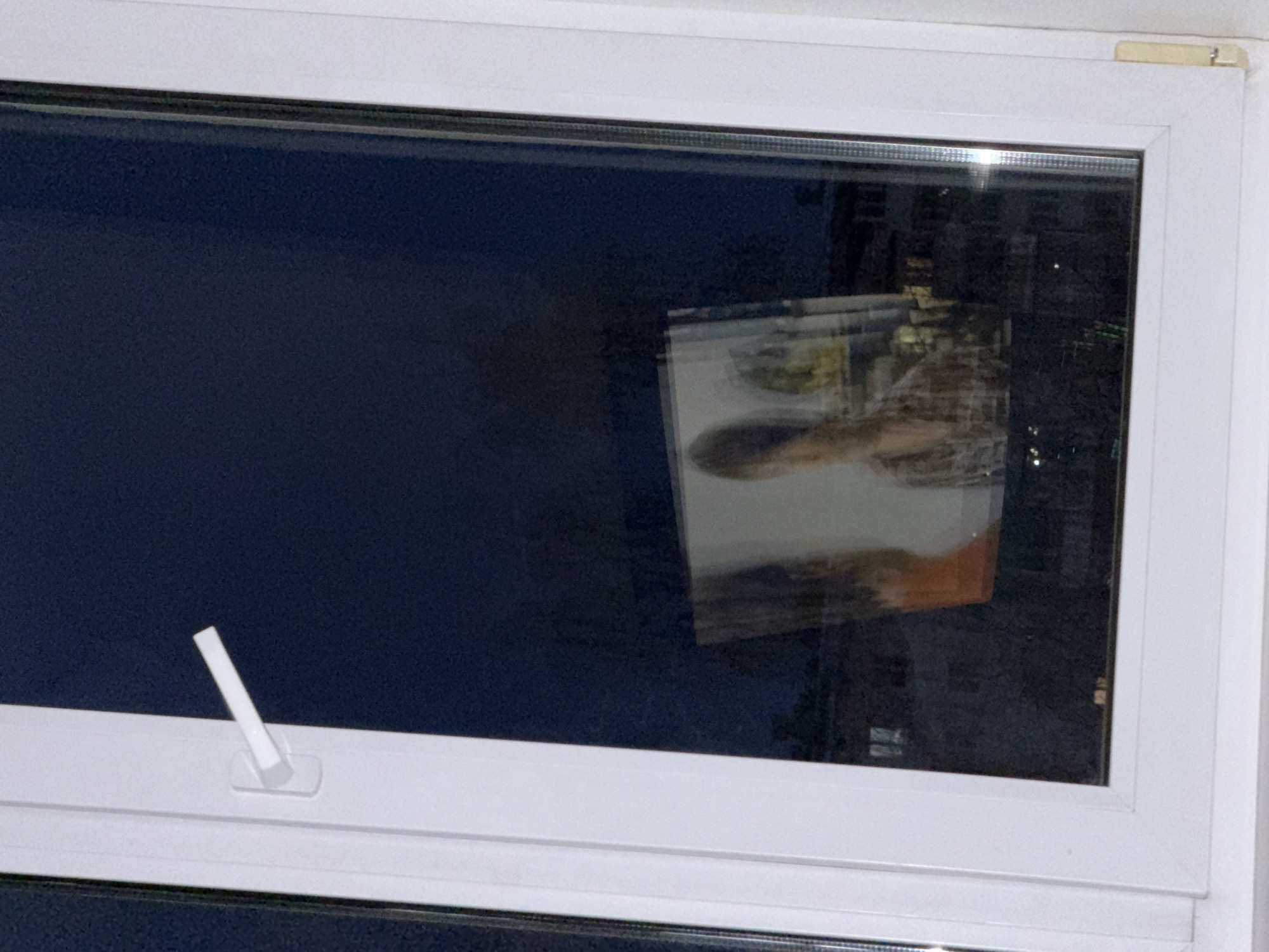 A white-framed window with a handle in the lower-left corner is seen. It appears to be nighttime, as the window shows a dark blue color. There is a reflection of an indoor scene, possibly a photograph, visible on the right side of the window. The city of Chaikovsky, Russia, is not discernible from the details in this image.