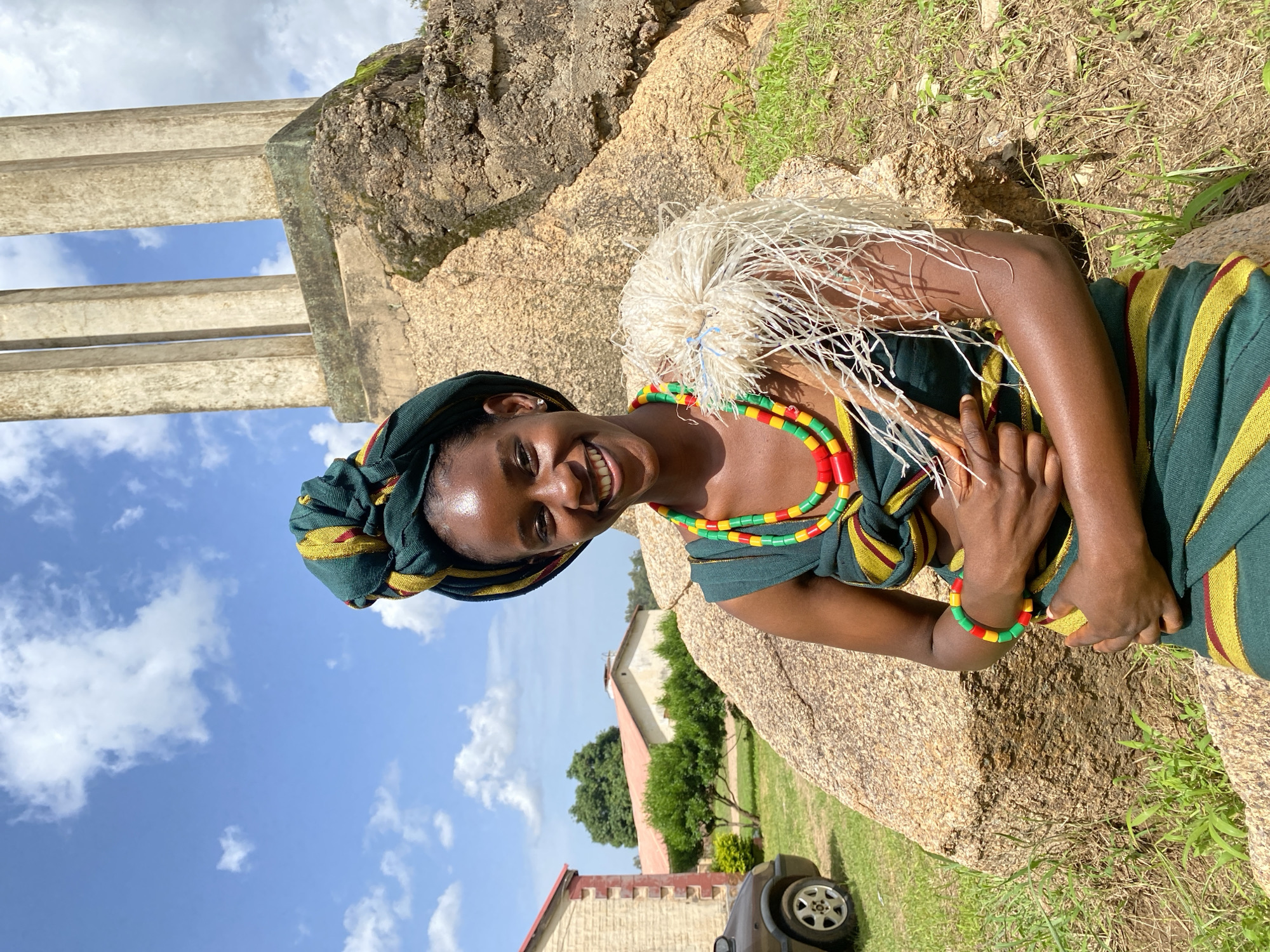 A young woman smiles broadly at the camera, leaning against a large, weathered rock. She wears a traditional green and yellow striped outfit with a matching headscarf. Around her neck and wrist are colorful beaded necklaces and bracelets in red, yellow, and green. A headdress made of white, straw-like material adorns her head. The scene appears to be outdoors, with a backdrop of large rocks and vegetation. In the distance, buildings with red roofs and a car can be seen. The sky is bright blue with scattered white clouds, suggesting it is daytime with good weather. The woman's expression is joyful and radiant. The image is a portrait, likely taken in Vom, Nigeria, given the context provided.