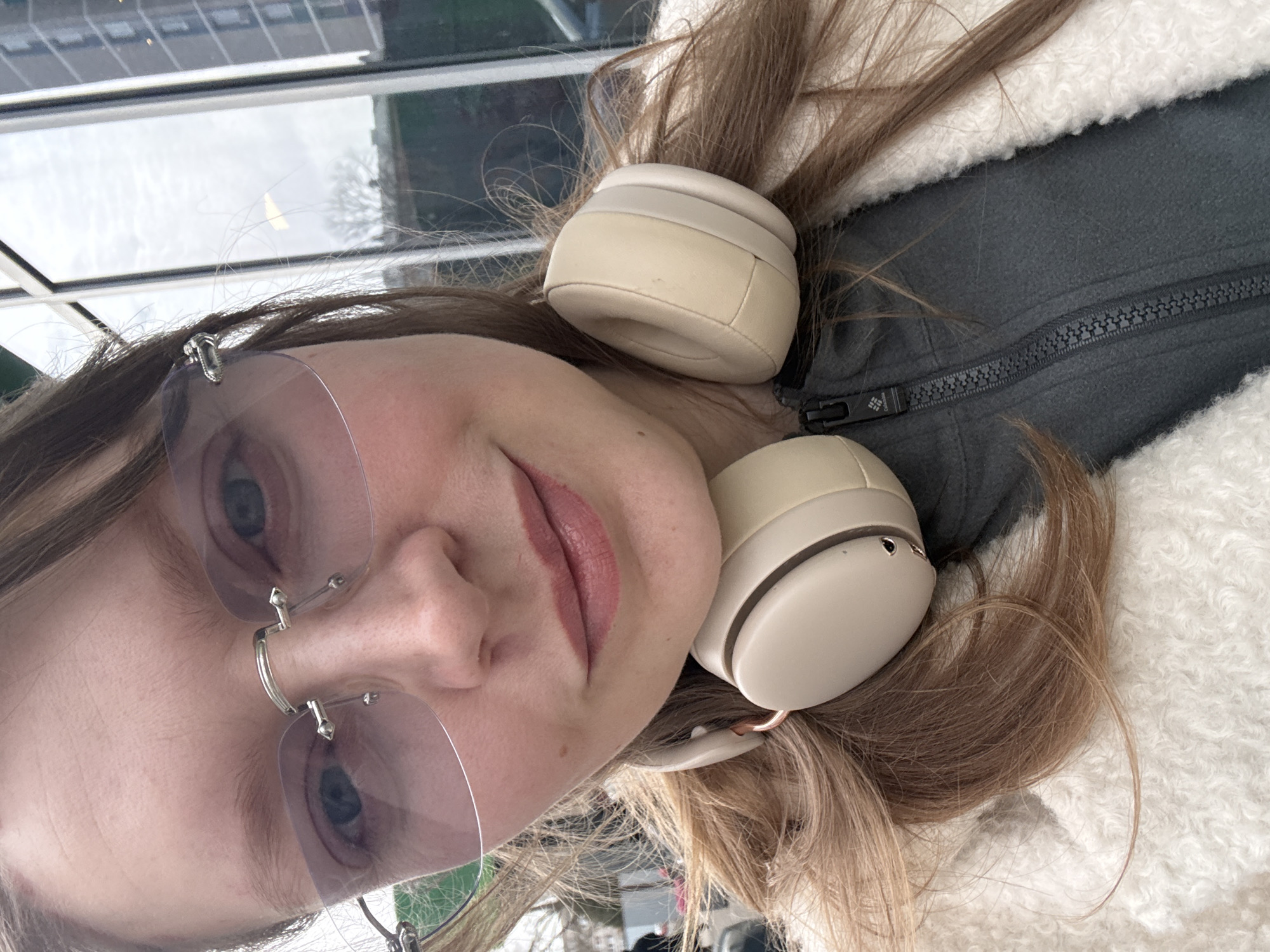 A medium shot depicts a woman with light skin, wearing wire-rimmed eyeglasses with a light tint, and light-colored headphones. Her hair is light brown with blonde highlights. She is wearing a white, fluffy jacket with a grey zipper. The background shows a blurry cityscape with a window frame, suggesting an indoor setting. The location in the city of Izhevsk, Russia is not discernible from the image.