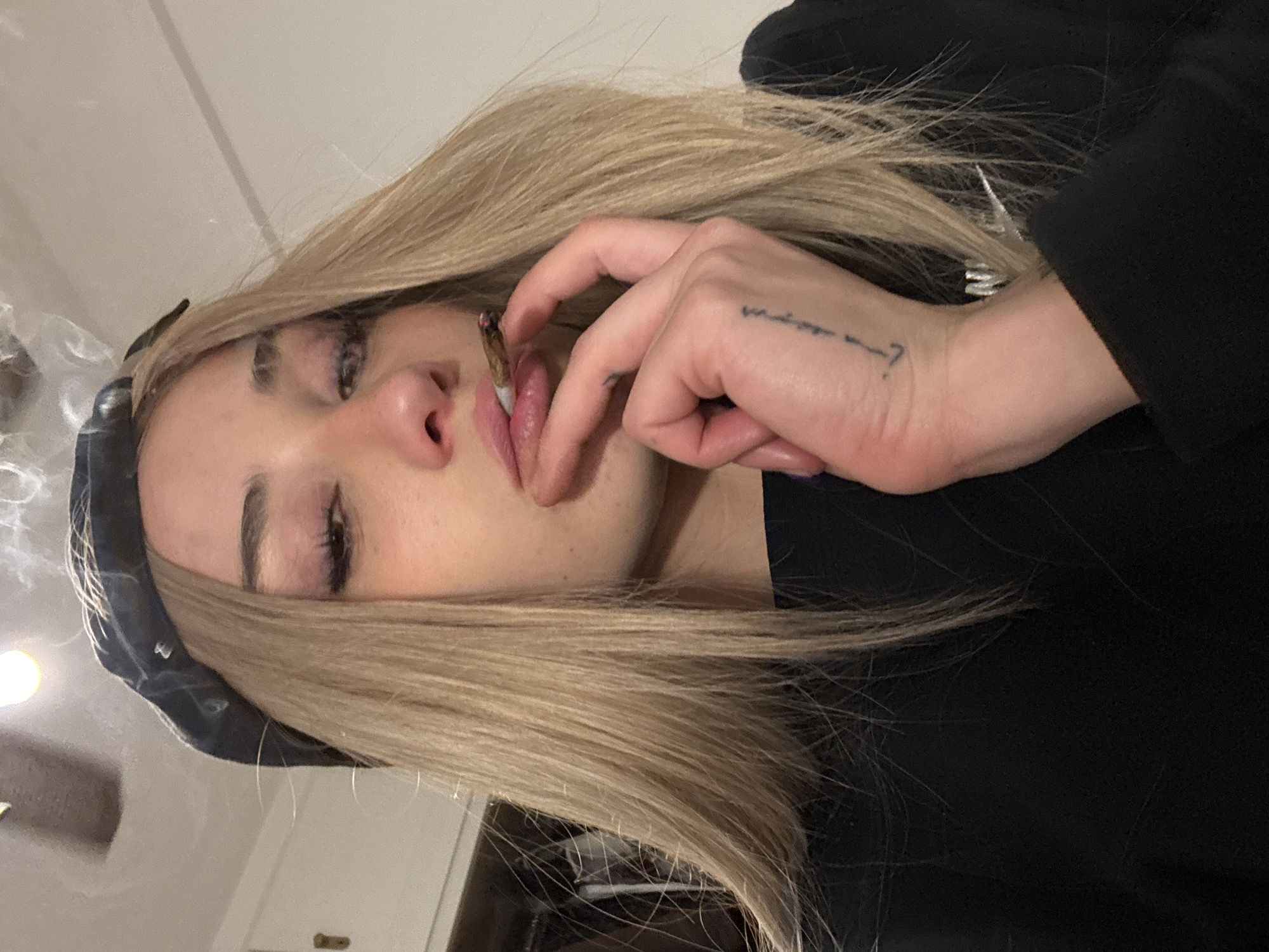 A fair-skinned person with blonde hair is holding a lit marijuana cigarette in their mouth with their left hand. They are wearing a black shirt and a black beret. The background is a blurred interior setting. The location is not determinable from the image.