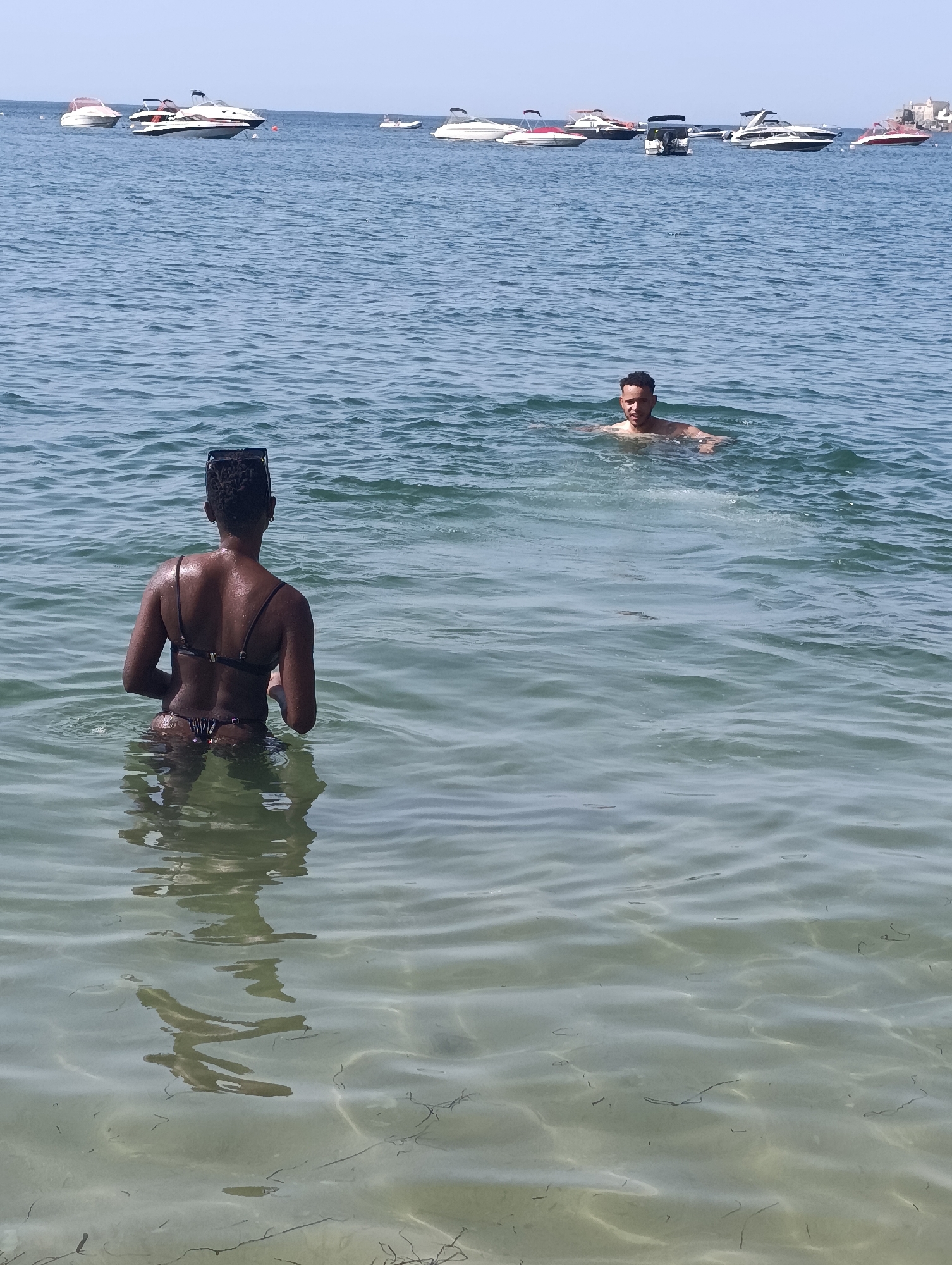 Two people are in the ocean near a coastline with several boats anchored. In the foreground, a Black person wearing a black bikini is facing away from the camera, standing in the shallow water. In the mid-ground, a light-skinned person is swimming and facing the camera. The water is clear, and the seabed is visible. The background shows a line of boats of various sizes, and in the far distance, a landmass with buildings is visible. The sky is clear and blue. The scene depicts a sunny day at a beach or bay.