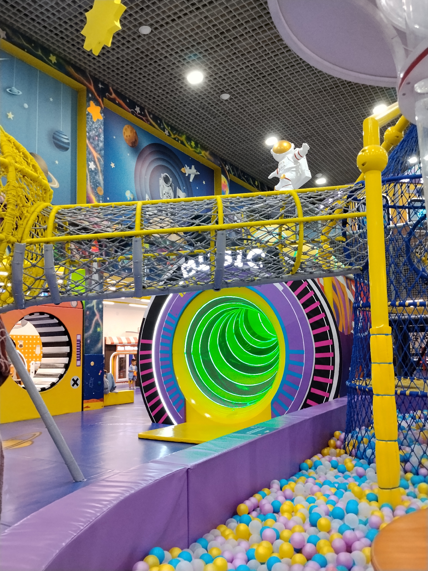 This is an interior shot of a children's play area with a space theme. The dominant colors are vibrant yellow, purple, and blue, with various circular and tunnel-like structures. In the background, a mural depicts a starry sky with planets and an astronaut. A plastic astronaut figure is suspended above a yellow net-enclosed walkway. A large, brightly lit green tunnel is visible in the center of the frame, surrounded by colorful, concentric circles. To the right, a yellow pole supports a blue net structure. In the foreground, a purple padded wall encloses a ball pit filled with multi-colored balls. A child is partially visible in the background near a small stall. The ceiling is a grid of acoustic tiles with recessed lighting. The floor is a smooth, purple mat. The location is identified as Bishkek City, Kyrgyzstan.