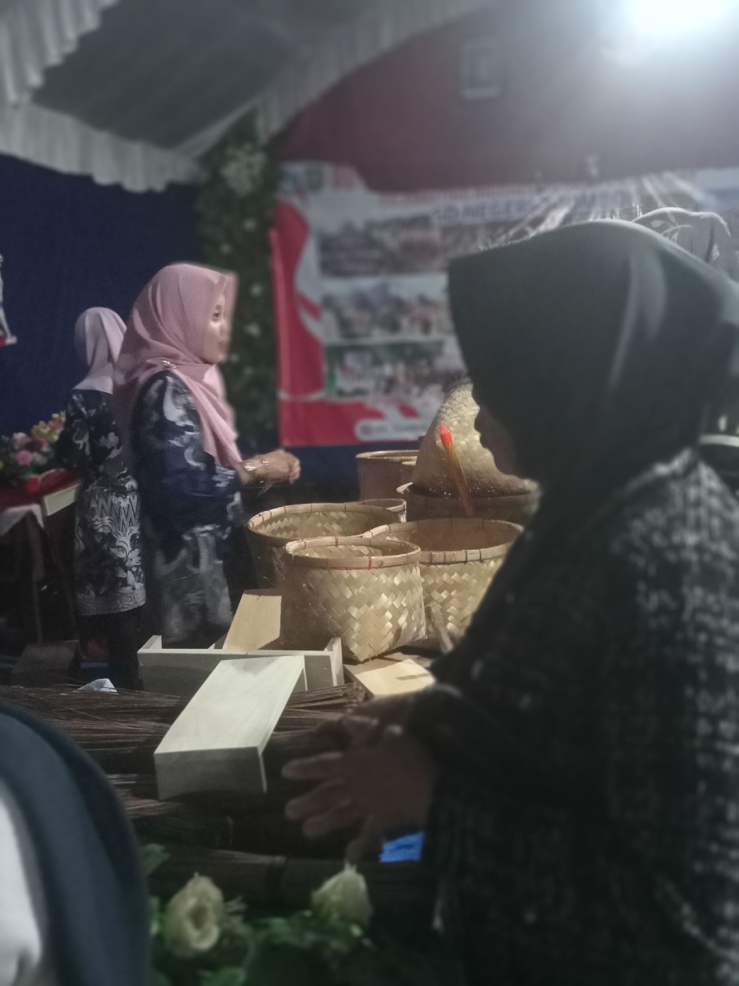 The image shows a dimly lit indoor scene, possibly a market or exhibition. In the foreground, a person wearing a dark headscarf and patterned clothing is partially visible, facing away from the camera. They appear to be interacting with objects on a table. In the middle ground, two women are standing, both wearing headscarves. The woman on the left is wearing a light pink headscarf and a dark patterned dress. The woman on the right is wearing a darker patterned dress. They are positioned near several woven baskets. The background features a dark blue curtain and a banner with text and images, suggesting a public event or display. The overall ambiance is that of a community gathering or fair in Pule, Indonesia.