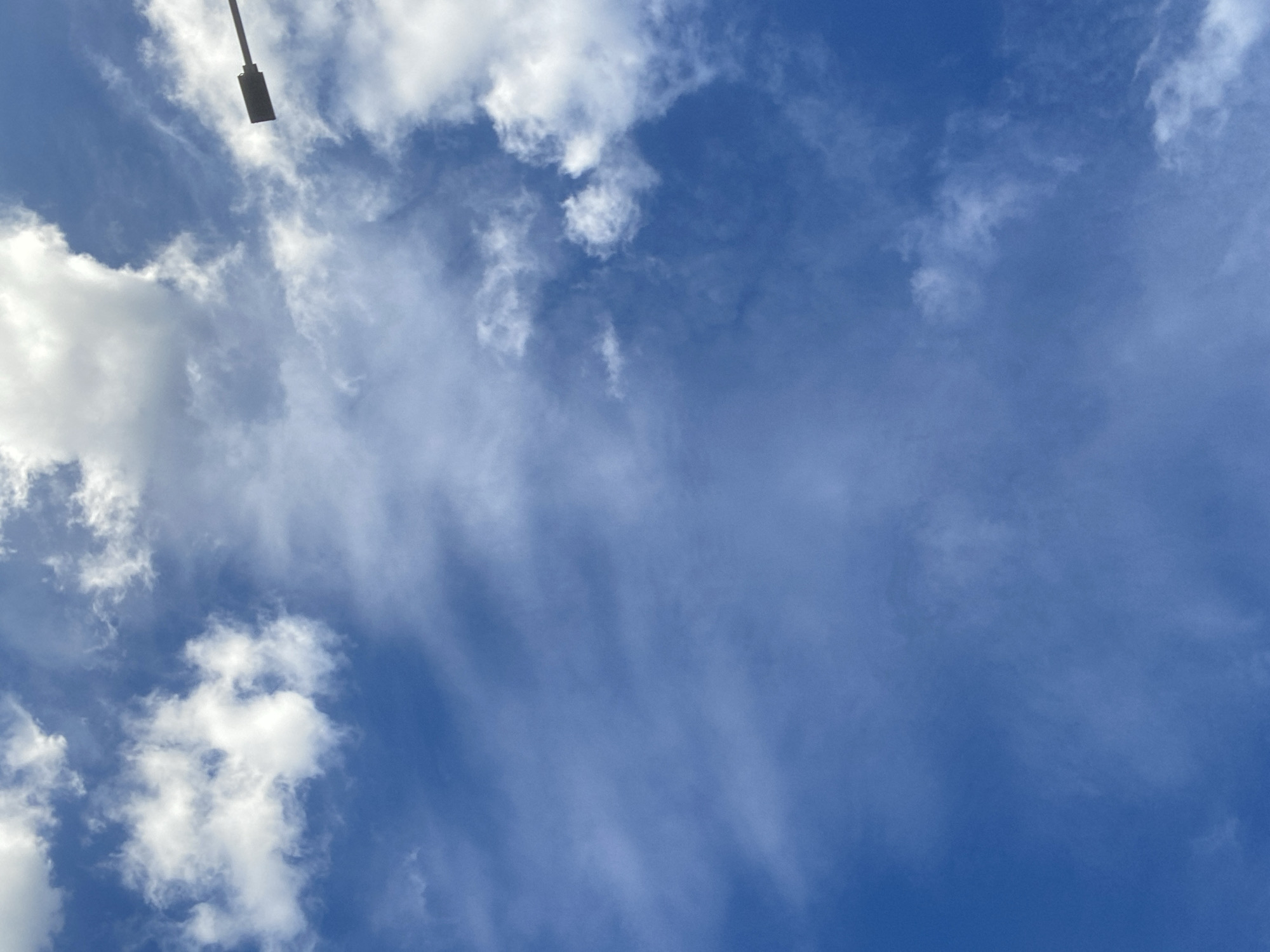 The image shows a clear blue sky with scattered white clouds. A dark, silhouetted street lamp arm extends from the top right corner of the frame. The scene is a sky-view, typical of an outdoor urban environment. The image does not contain any information to place it in Chaikovsky, Russia.