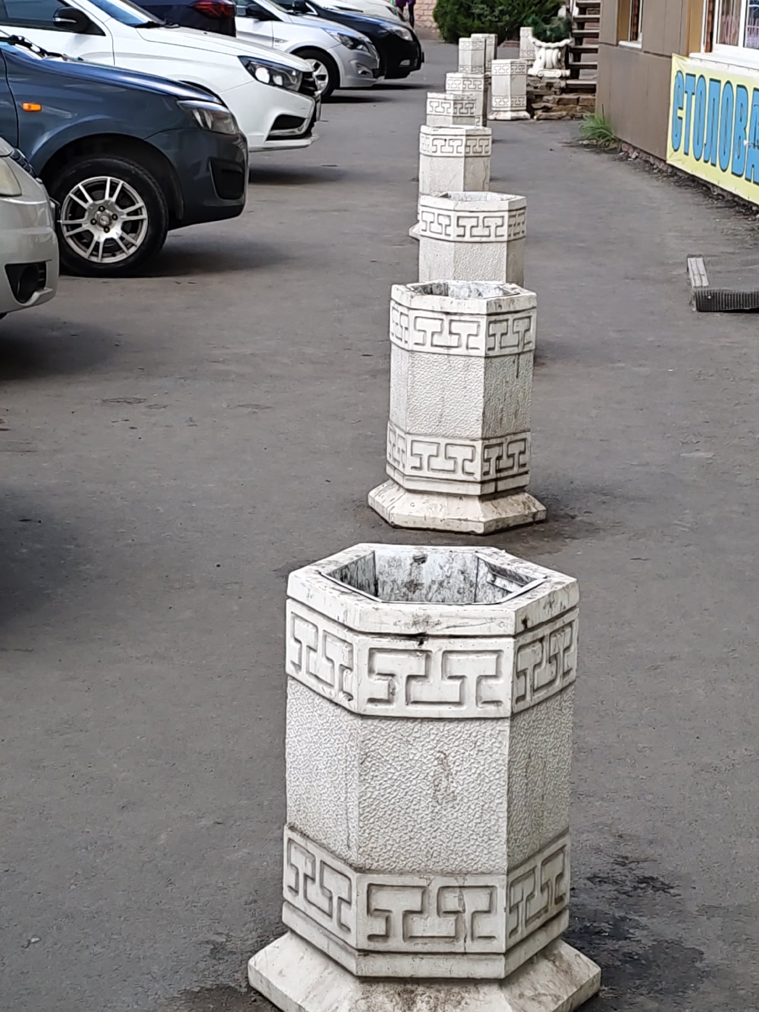 A row of ornate, hexagonal concrete planters with a Greek key pattern lines a paved area. The planters are weathered and some appear to be filled with debris. In the background, cars are parked and a building with a yellow sign is visible. The scene is likely in Rostov-on-Don, Russia, based on the presence of the planters which are common in the city.