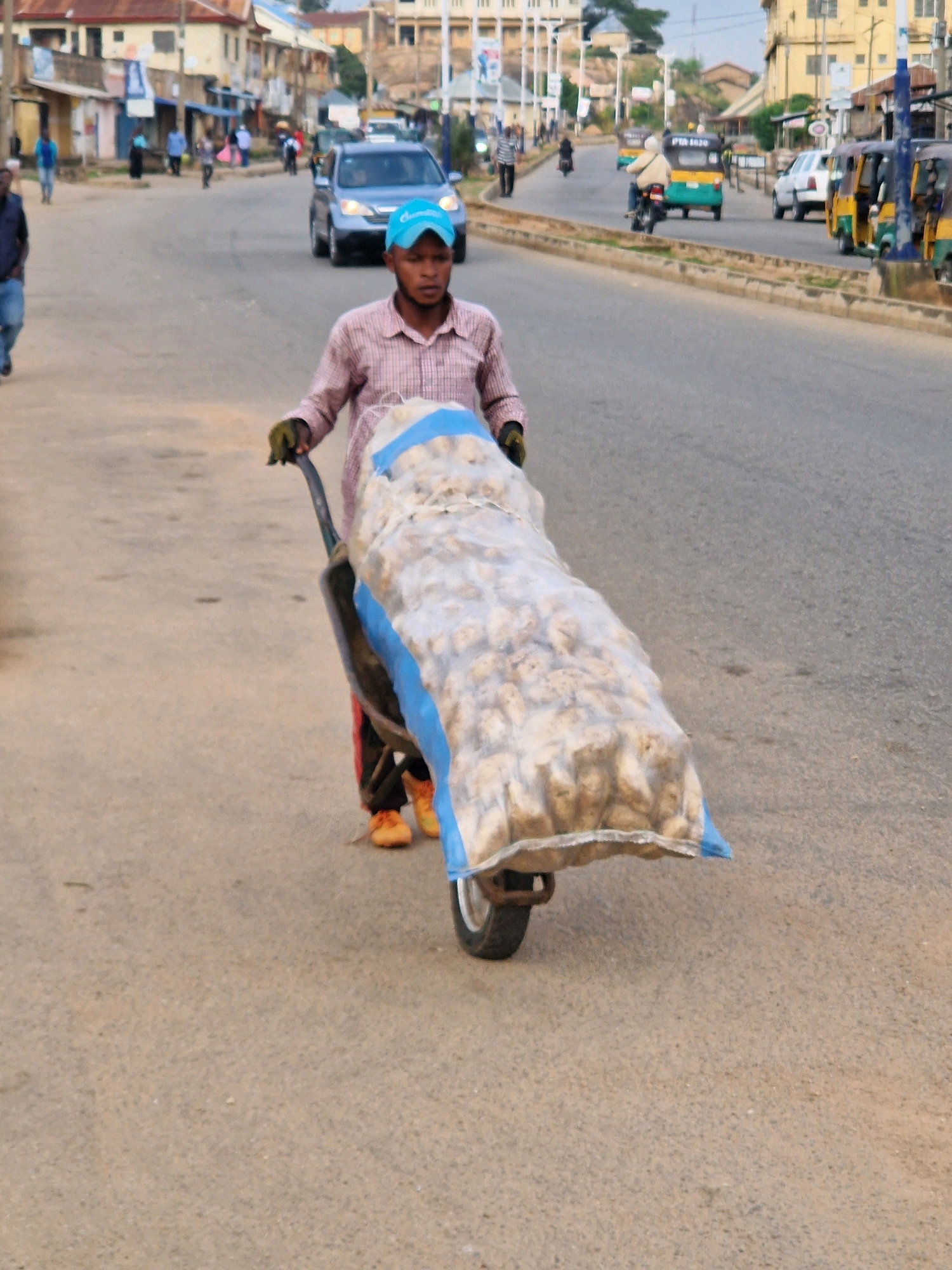 A man, wearing a pink checkered shirt, a blue baseball cap, and orange shoes, is pushing a wheelbarrow full of bagged goods down a paved road. The wheelbarrow is laden with a large, clear plastic bag filled with what appear to be potatoes. The man is holding the handles of the wheelbarrow, his body turned to the side as he propels it forward. The road is lined with buildings and businesses on either side, and other vehicles, including cars and motorcycles, are visible in the background. The scene appears to be in an urban or semi-urban area, possibly in Kabong, Nigeria, given the architectural style and types of vehicles. The sky is overcast.