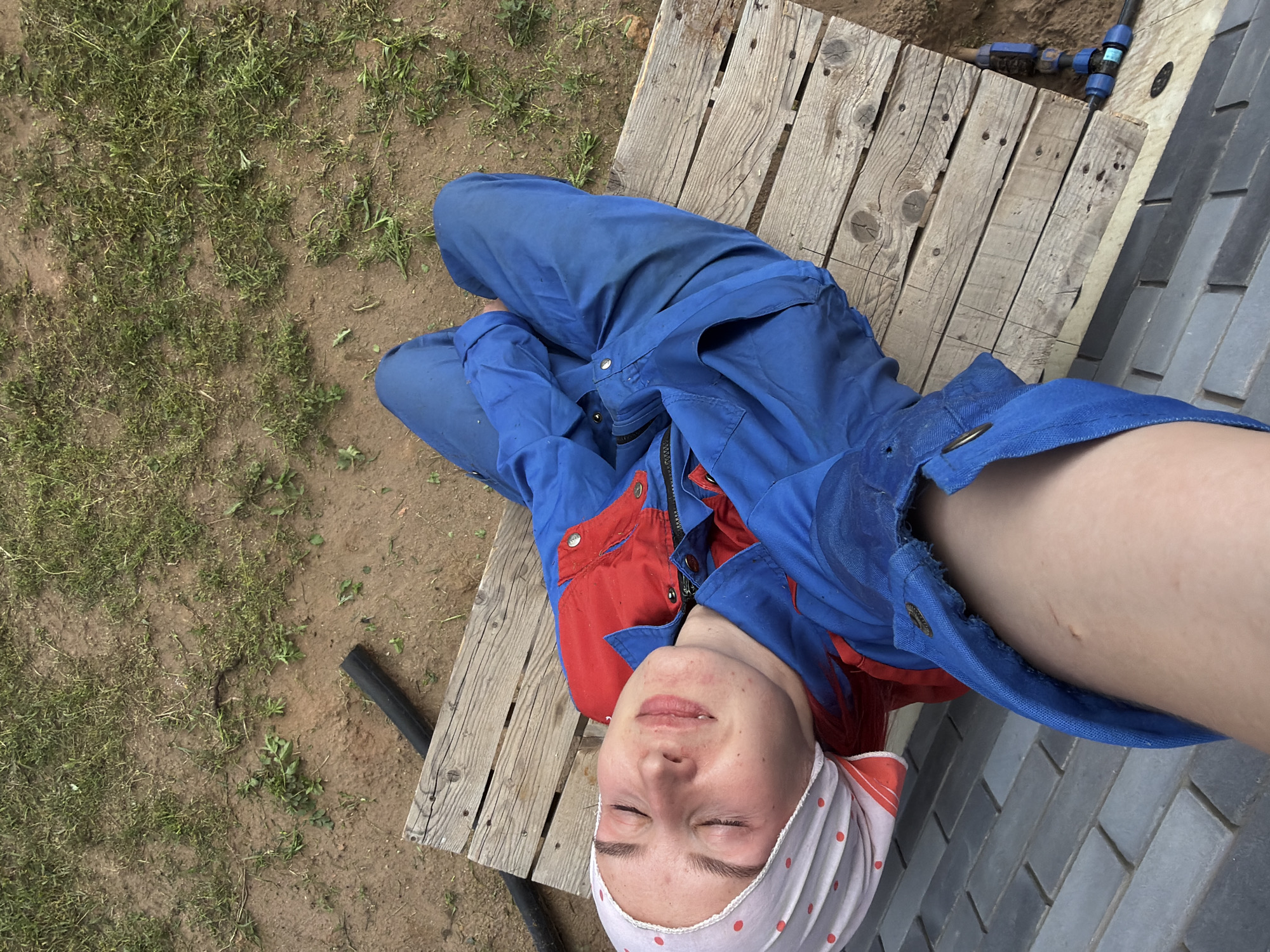 A young person in blue overalls with red accents and a red and white polka dot headscarf is lying on a wooden pallet outdoors. The person's eyes are closed, and they appear to be relaxed. The ground around the pallet is dirt with sparse grass. A black pipe is visible to the left of the pallet, and a blue irrigation system is attached to the right. The overall scene suggests an outdoor work or leisure activity. There is no indication of the city of Chaikovsky, Russia.