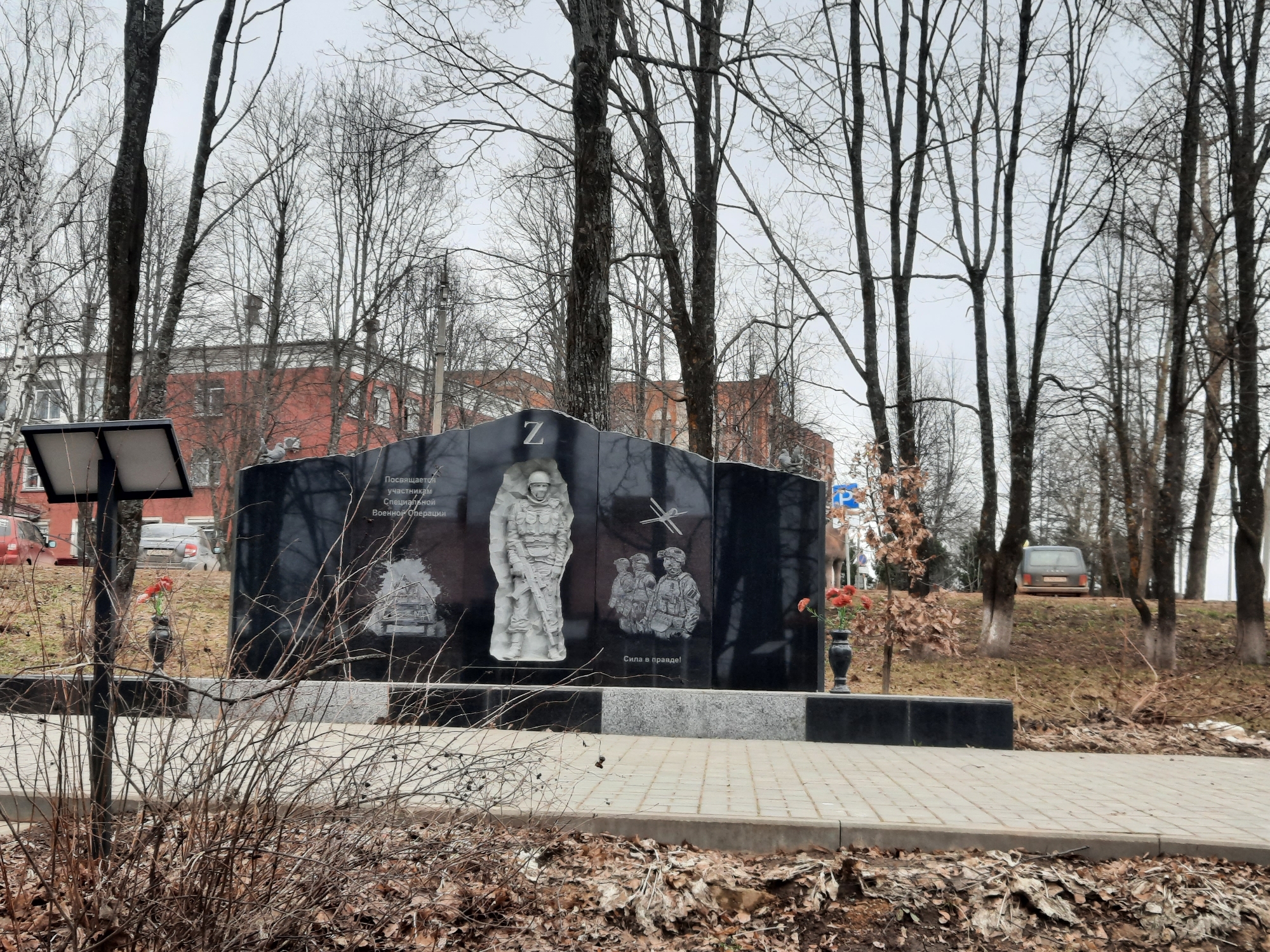 The image shows a monument dedicated to participants of the "Special Military Operation" in the city of Krasnogorskoye, Russia. The monument is made of black polished stone, featuring engraved depictions of soldiers, military equipment (including an airplane), and the letter "Z," a symbol associated with the Russian military campaign in Ukraine. Text in Russian is also visible on the monument.
The monument is located outdoors in a park-like setting. Bare trees surround the area, indicating it is likely late autumn or winter. There is a paved pathway leading to the monument. A bush in the foreground is bare. In the background, there is a building with red brick walls. Cars are visible parked in the vicinity. A gray sedan is parked near the edge of a hill. The weather appears to be overcast. Flowers have been placed at the base of the monument.