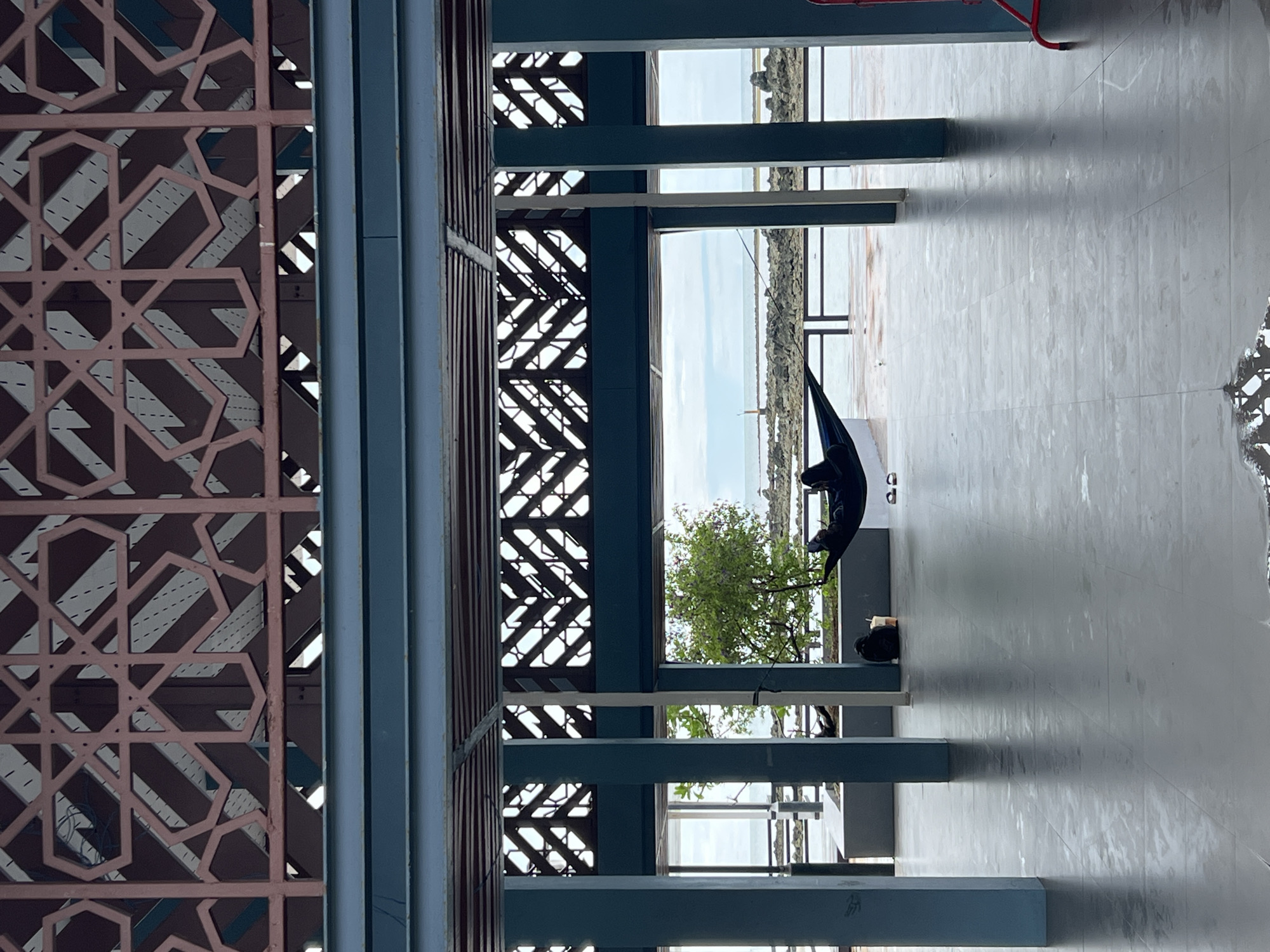 The image depicts an outdoor structure in Baan Hua Thanon, Thailand. The structure features ornate, patterned screens and blue-painted columns and beams. A tree and a body of water are visible through the openings of the structure. On the tiled floor within the structure, a person dressed in black is present, seemingly seated or lying down. The scene suggests a modern design with traditional influences, situated near a waterfront location.