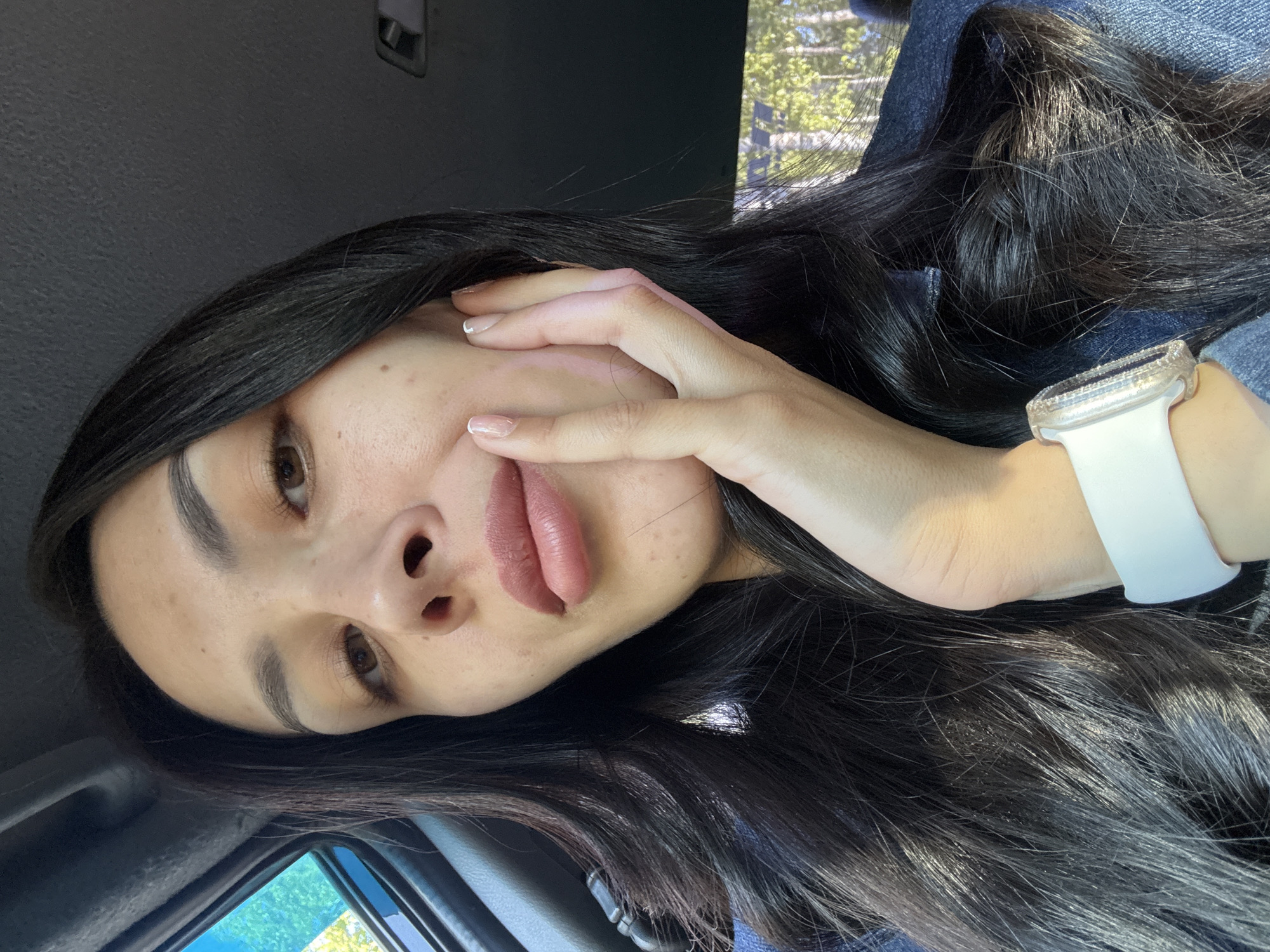 A young woman with long, dark hair is in a car. She is wearing a white smartwatch on her left wrist. She is holding her left hand to her face. Her lips are mauve. The background visible through the car window suggests a location with trees and possibly buildings in the distance. It's not possible to confirm the location in the city of Votkinsk, Russia with the given information.