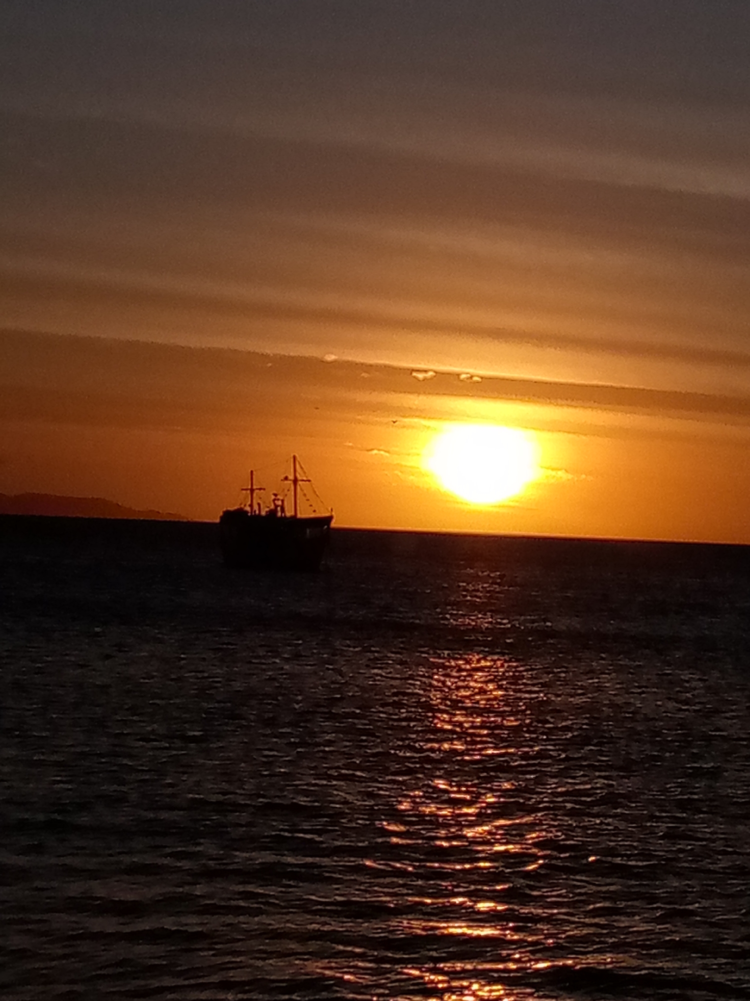 A wide shot captures a fishing vessel against a backdrop of a golden sunset over the ocean. The silhouette of the boat, featuring a mast and rigging, is positioned near the center of the frame, slightly to the left. The sun, a bright disc, sits just above the horizon, casting a shimmering reflection on the water. The sky transitions from dark hues above to warm orange tones closer to the horizon. The environment is dark, with light reflecting on the water. This scene depicts a sunset in Juan Griego, Venezuela.