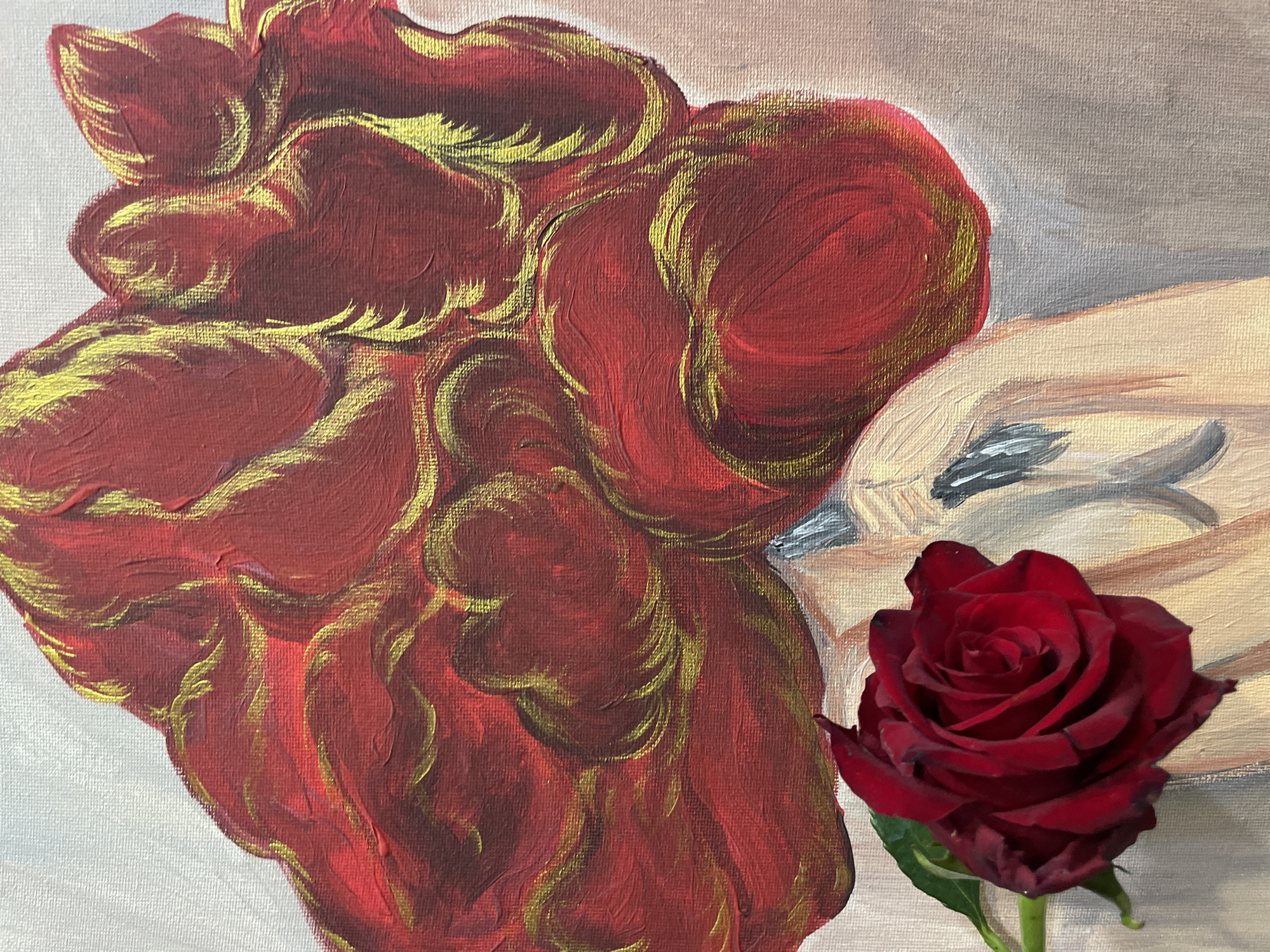 A close-up image shows a partially painted canvas with a real rose placed in front of it. The painted section of the canvas depicts a red and gold flower or design to the left, with a light-skinned face visible to the right of the flower. The face is in profile with closed eyes, appearing to be asleep or at rest. A real, dark red rose with a green stem is situated in the foreground to the right, in front of the painted image. The location of the image is not discernible from the picture.