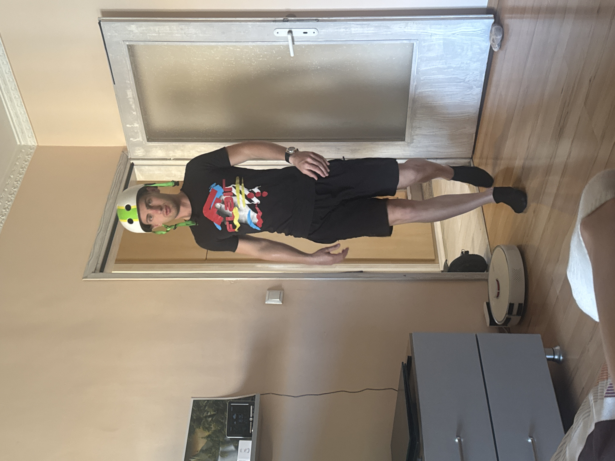 A man wearing a white and green helmet, a black t-shirt with a colorful graphic, and black shorts walks towards a glass-paned door. He is also wearing black socks. A white robotic vacuum cleaner is on the wooden floor to his left, and a gray dresser is against the wall to his far left. Above the dresser, a shelf holds a framed picture and a digital clock. The scene is indoors, likely a residential setting.