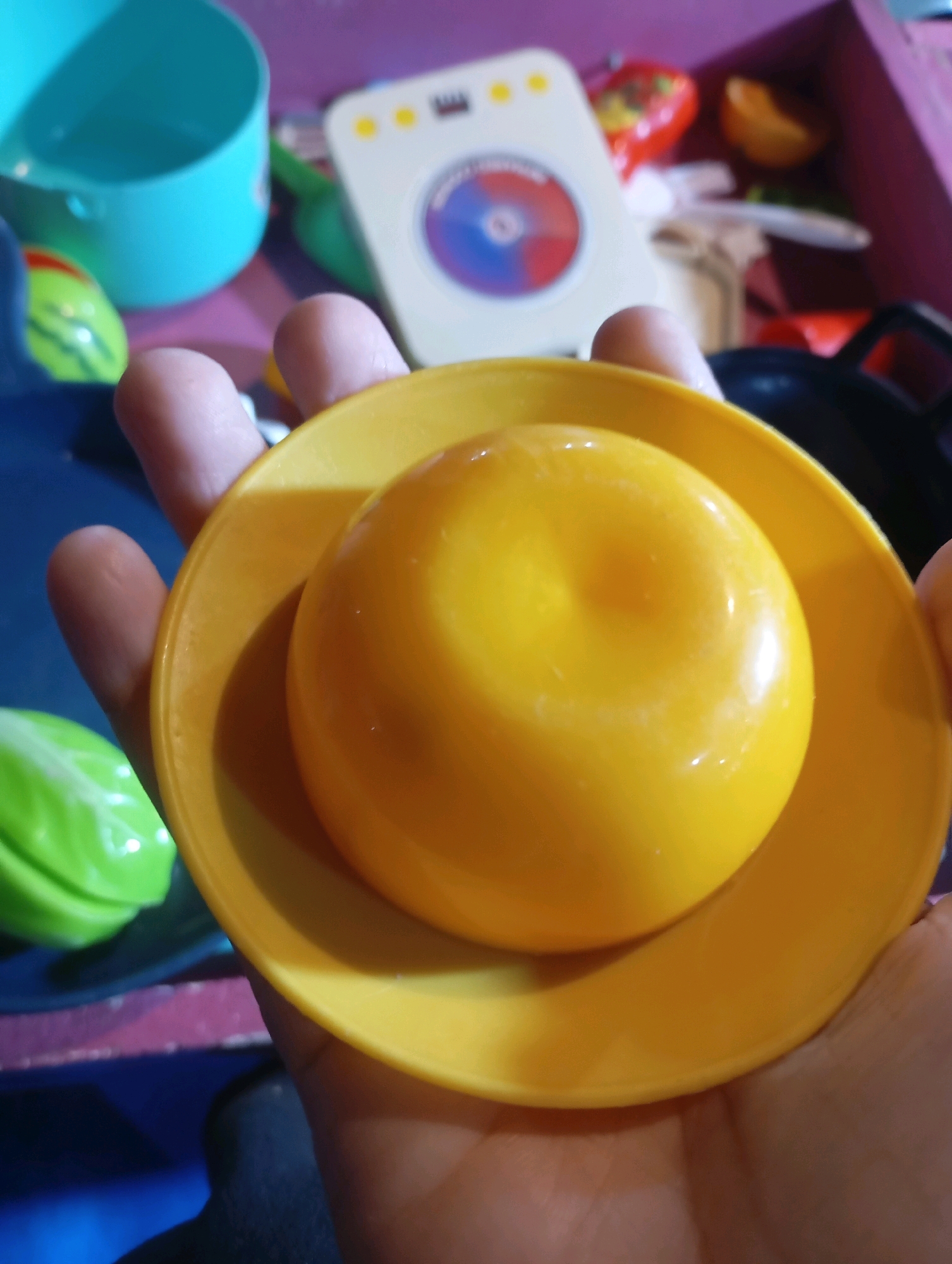 A close-up, top-down shot shows a person's hand holding a yellow toy play food item, possibly an apple, on a matching yellow toy plate. The toy is shiny and appears to be plastic. The background is blurred and shows other toy items, including a green toy that resembles lettuce, a turquoise bucket, and what looks like a toy washing machine. The scene is dimly lit, with some reflections visible on the toys. The overall impression is that of a child's play area. This image does not provide any information about the city of Randegan, Indonesia.