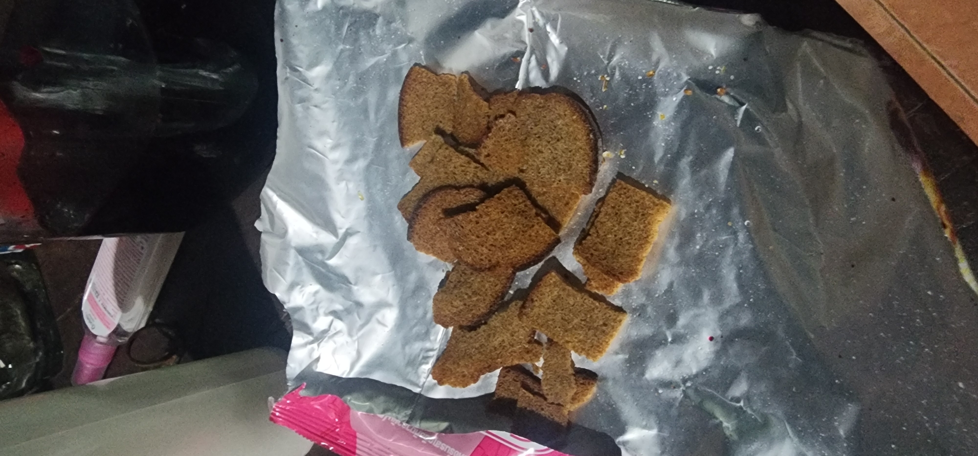 The image displays a pile of sliced rye bread, likely croutons or rusks, arranged on a sheet of crumpled aluminum foil. The bread is a dark brown color with a coarse texture, and some pieces are broken. The foil is shiny and reflects light. In the background, parts of other objects are visible, including what appears to be a bottle with dark liquid and a pink and white spray bottle. A portion of a pink and white packet is also visible on the left side of the frame. The overall scene suggests a casual setting, possibly an outdoor picnic or a preparation area for food. There is no discernible indication of a specific location within the city of Baan Bo Phut, Thailand.