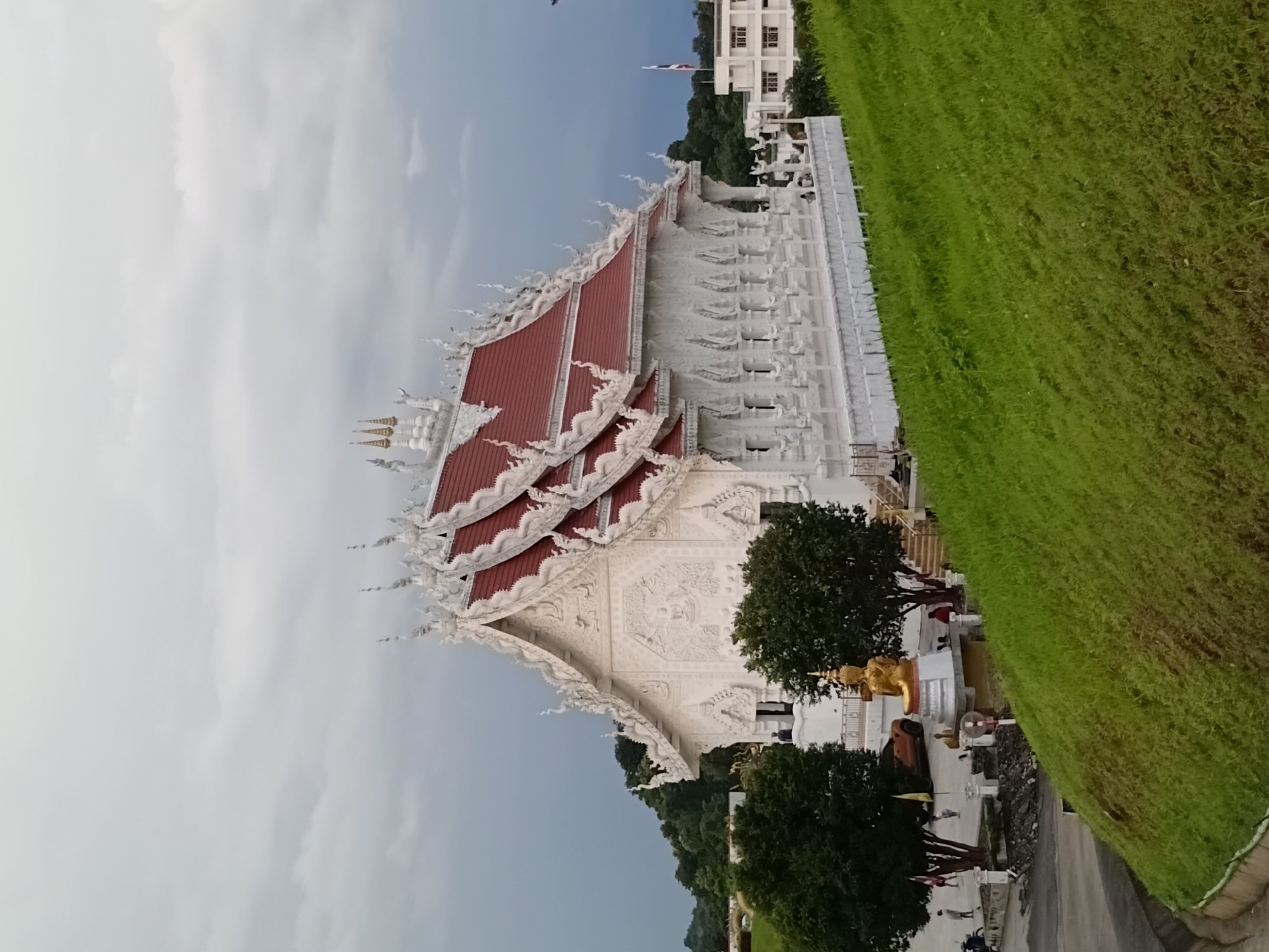 The image shows a white temple with red roofing, featuring intricate carvings and ornate details. A golden Buddha statue is positioned in front of the temple. The temple is surrounded by lush green trees and a grassy hill. In the background, there is a modern building and a flagpole. The sky is overcast. The scene is located in Ban Lao Phatthana, Thailand. There are several people visible in the image, some standing and observing the temple, while others are near a vehicle. The overall impression is of a peaceful and spiritual setting.
