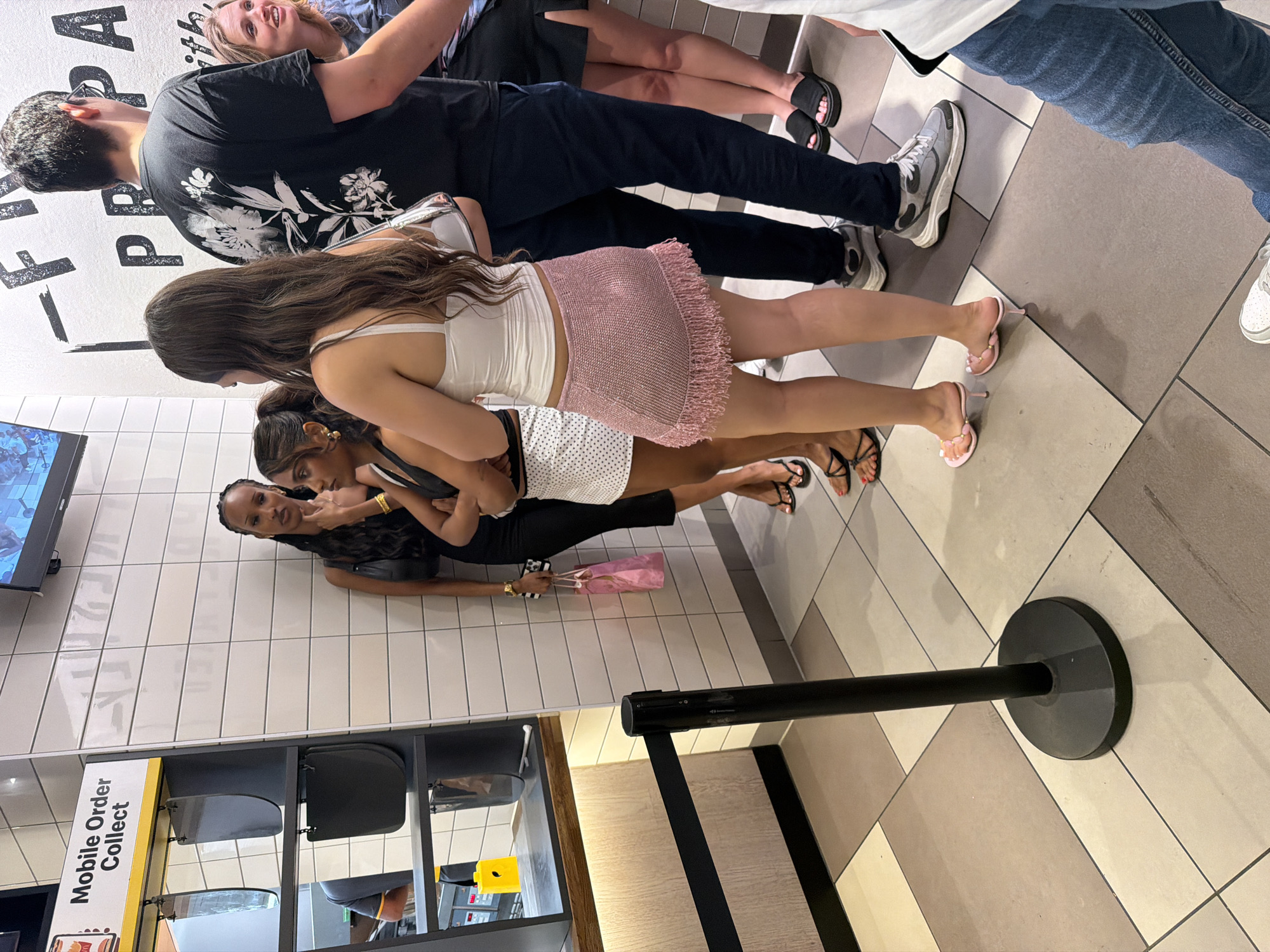 The image shows a queue of people in a McDonald's restaurant. A "Mobile Order Collect" sign is visible near a shelving unit with various items. Several people are standing in line, and a black stanchion with a retractable belt is used for queue management. The restaurant has a tiled floor and a tiled wall. An electronic display is visible on the wall. The location is likely within the city of London, United Kingdom, given the presence of a McDonald's and the style of the signage and queuing system.