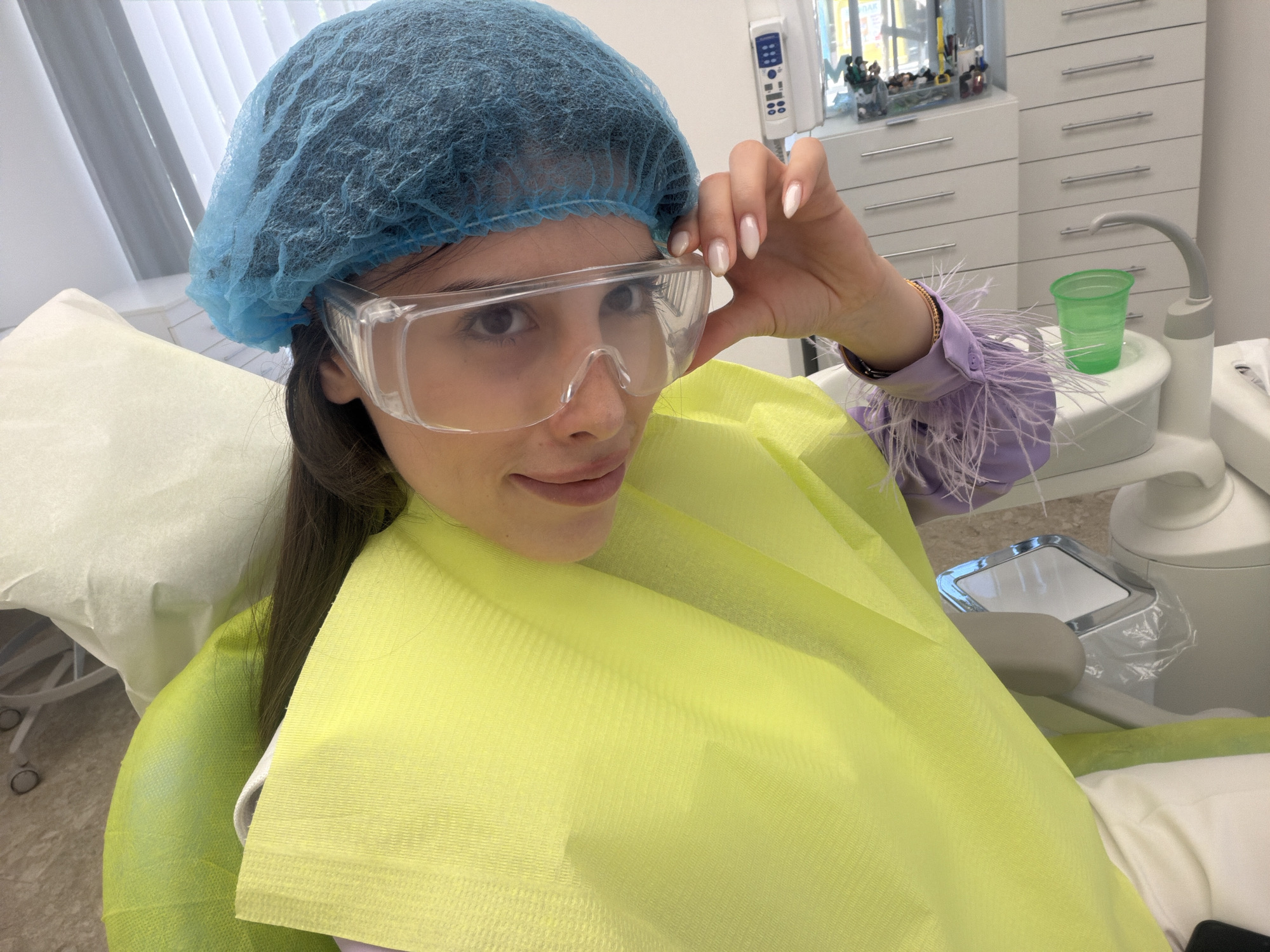A woman is seated in a dental chair, wearing a blue surgical cap and clear protective goggles. She has a neon yellow dental bib draped over her chest and shoulders. She is looking at the camera and adjusting her goggles with her right hand, which has neatly manicured nails. She is wearing a purple shirt with feather accents on the sleeves. The background shows a dental office environment with a white cabinet filled with drawers and a green plastic cup on a dental unit. A white headrest and a portion of the dental chair are also visible.