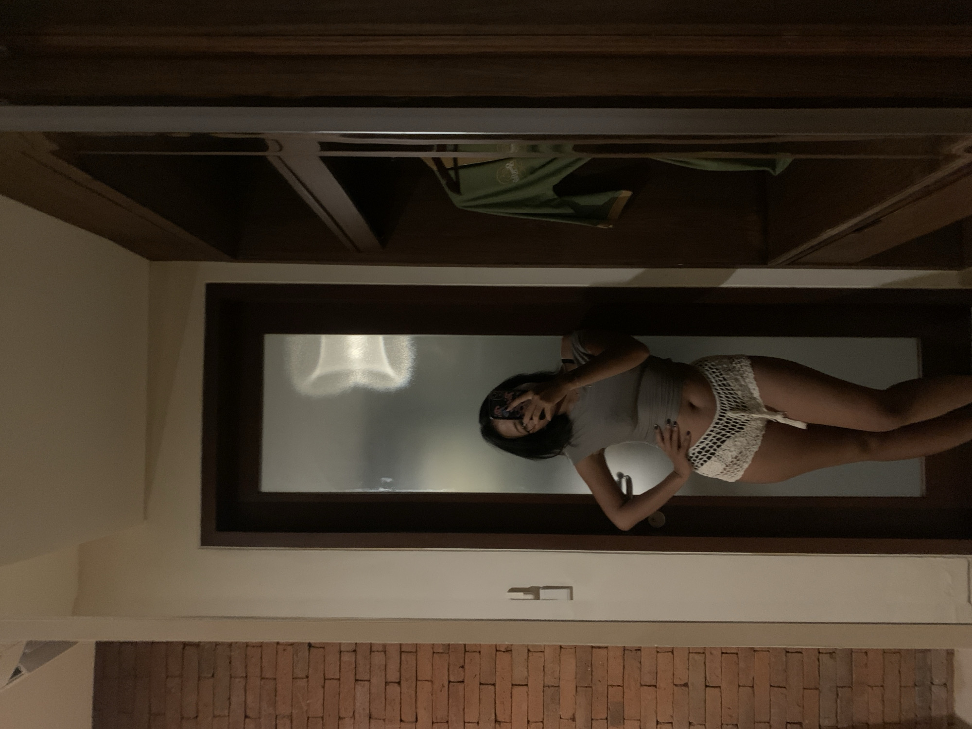 The image shows a person taking a selfie in what appears to be a bathroom. The person is wearing a gray top and white crocheted shorts. They are holding a phone in front of their face, partially obscuring it. The background features a window with frosted glass, a white wall, and a brick wall. Above the person is a wooden ceiling or shelf. The location is identified as being in the Ubud District, Indonesia.