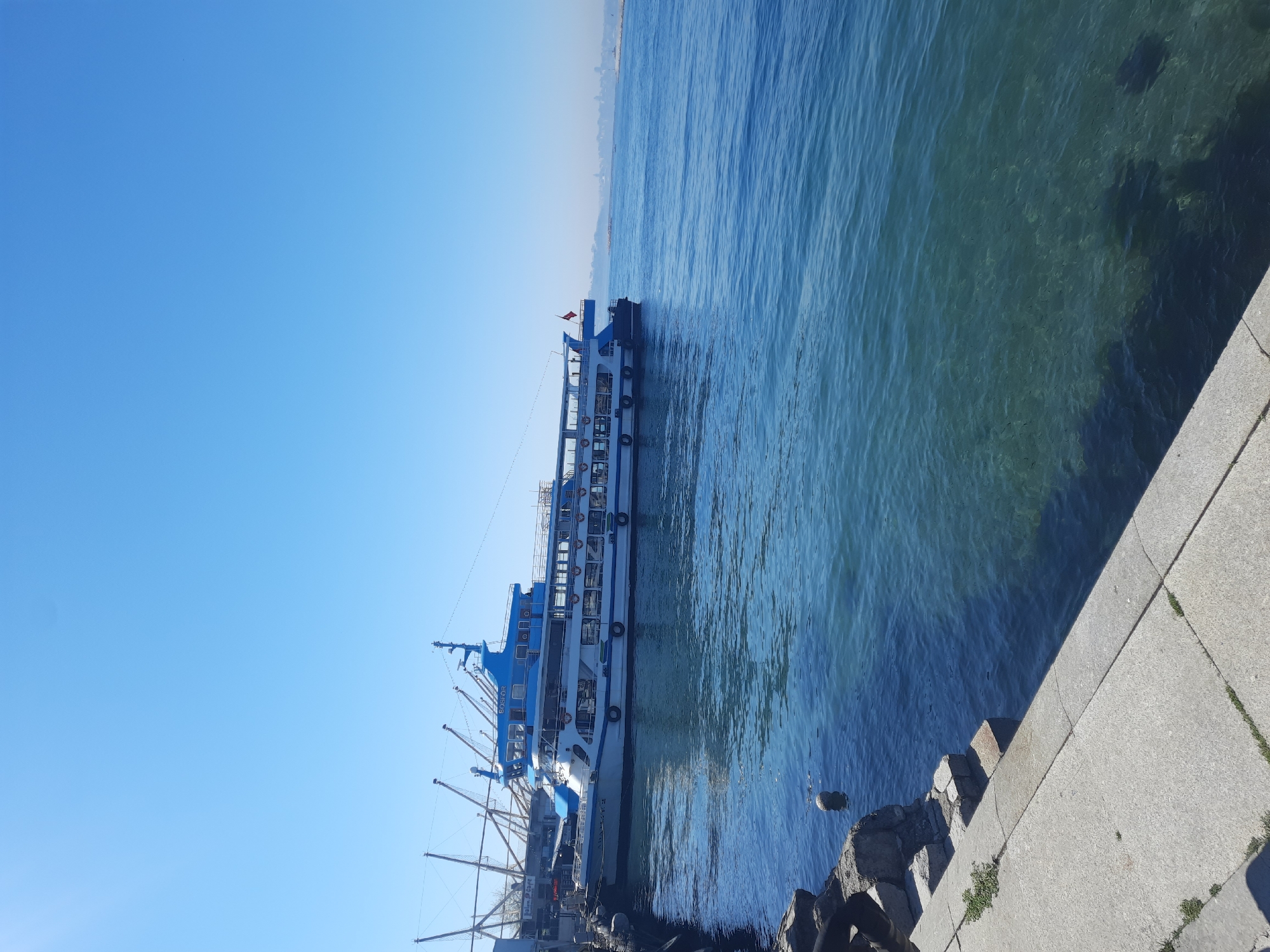 A high-angle shot depicts a large, white passenger ferry with blue trim docked at a concrete and stone embankment in Kadıköy, Turkey. The vessel is parallel to the dock with mooring lines visible. The blue water shows some transparency revealing the sea floor, and beyond the boat stretches to the horizon where a faint cityscape is visible. The sky is clear and light blue.