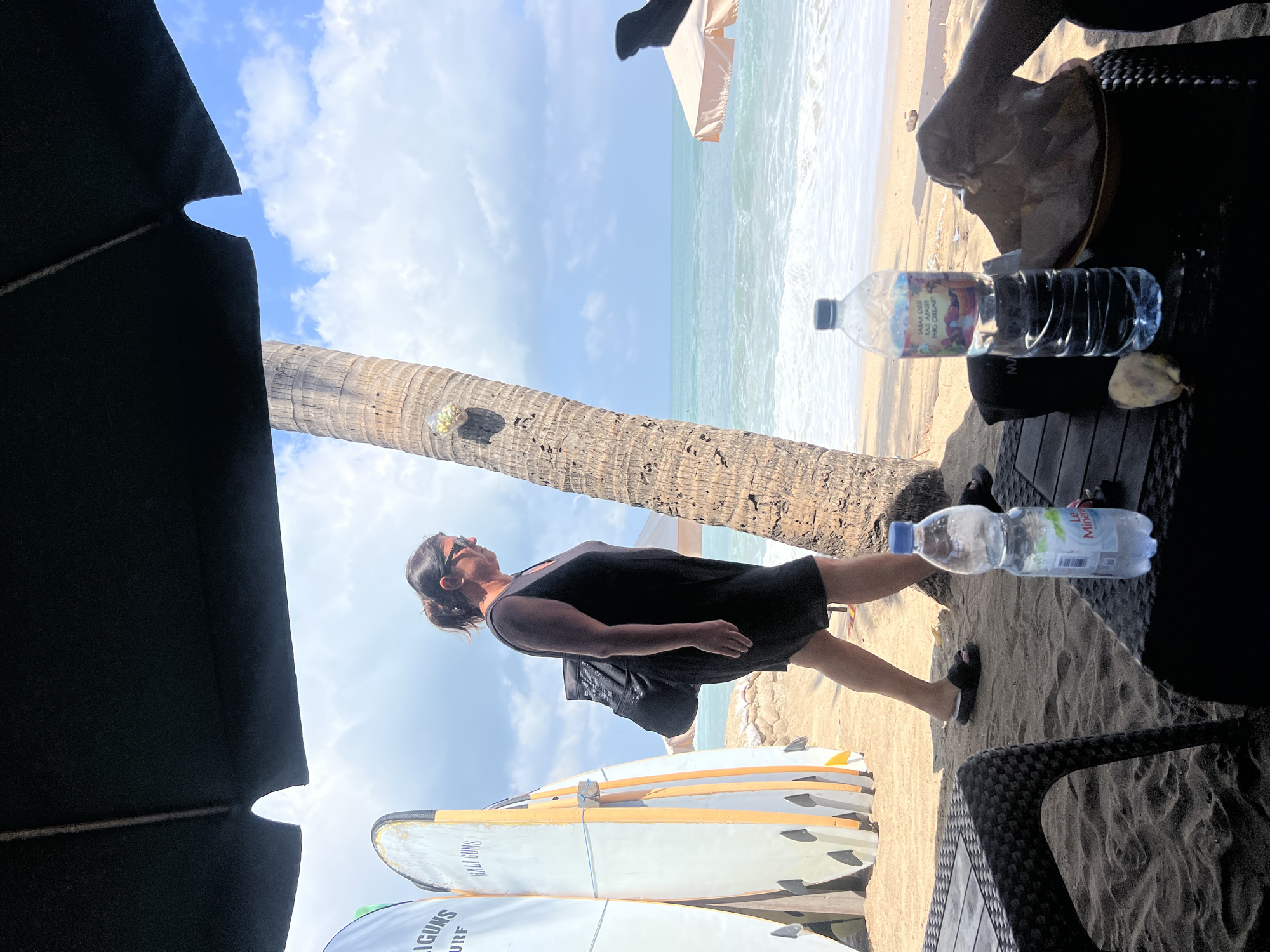 The image depicts a beach scene in Kuta, Indonesia, under a large black umbrella. In the foreground, two woven tables are set on the sand. On the table to the right, a person's arm is visible, holding a plate of food and a bottle of water. Another bottle of water rests on the table to the left. Behind the tables, a woman in a black dress walks along the sandy beach, carrying a backpack. She is walking towards the right of the frame, with surfboards lined up to her left. Further in the background, the ocean waves crash onto the shore under a partly cloudy blue sky, and a small beige umbrella is visible. A palm tree stands in the middle ground, its trunk marked with some objects attached to it.