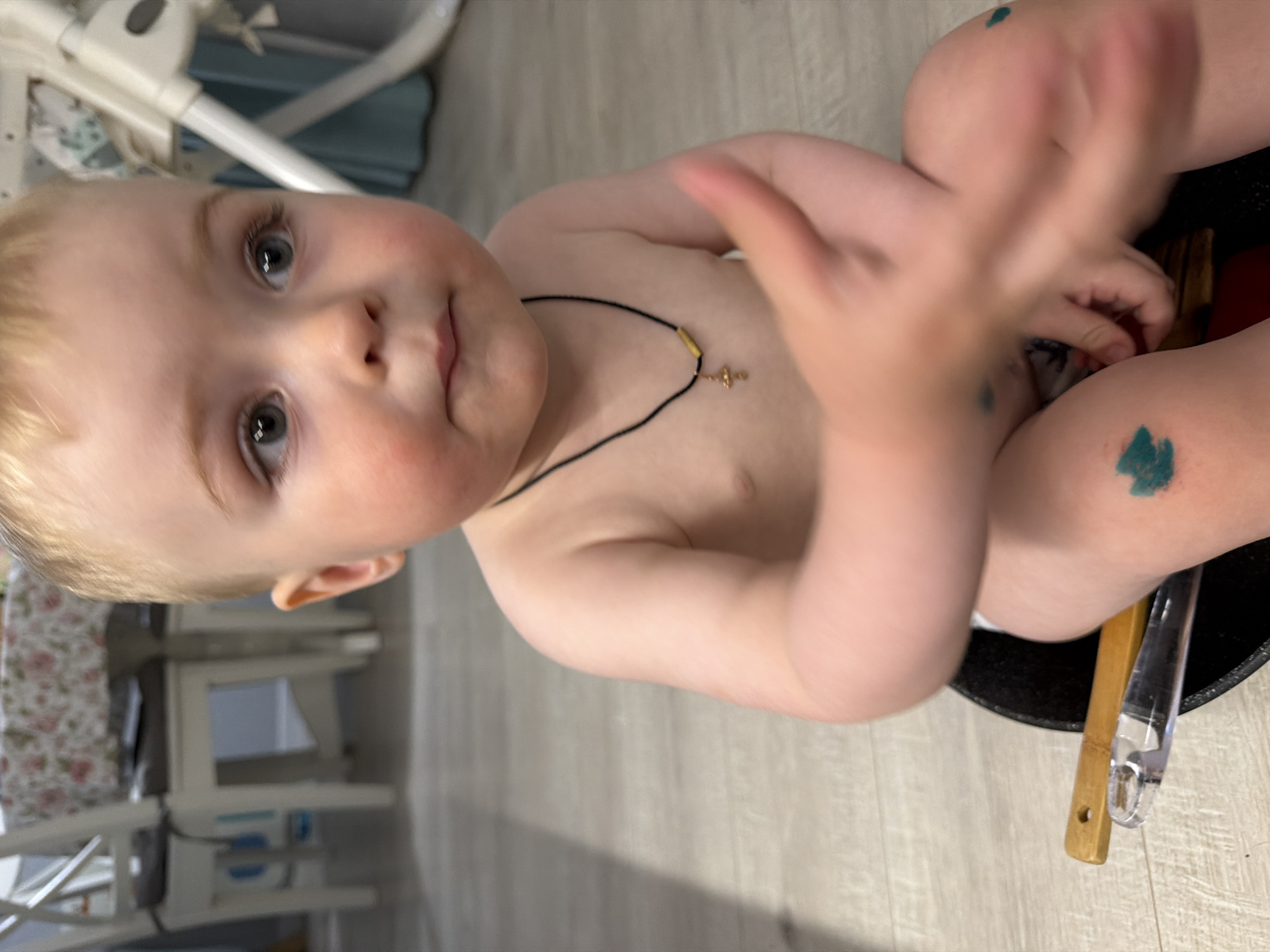 A baby is sitting indoors. The baby has blonde hair and blue eyes, with small dabs of green paint on its knees. The baby is wearing a necklace with a small cross pendant. The baby is sitting in a black container or bucket, with two utensils, one wooden and one plastic, placed inside. In the background are furniture items including a high chair and dining chairs. There is no information available to determine the location is in Votkinsk, Russia.