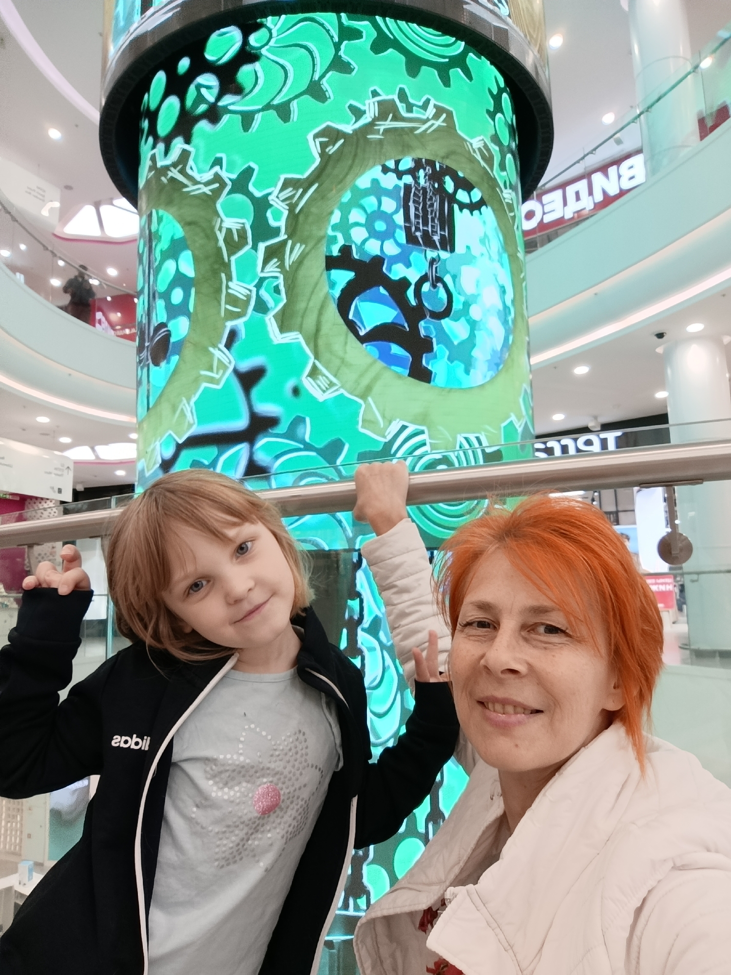 Here is a detailed description of the image:
**Scene:**
The image captures a scene inside a modern shopping mall. A large cylindrical pillar dominates the center of the shot, adorned with a digital display featuring intricate clockwork and gear designs in shades of green and blue. The mall's interior exhibits multiple levels with glass railings, visible in the background. Stores and signage can be discerned on the upper levels.
**People:**
Two individuals are the primary focus: a young girl and an adult woman. The girl has fair skin, light brown hair, and is wearing a dark-colored jacket over a light-colored t-shirt with a circular design. She is looking directly at the camera with a slight smile.
The woman has fair skin and short, bright red hair. She is wearing a light-colored jacket and also faces the camera, smiling. They are close together, suggesting a familial relationship, possibly mother and daughter. They are both holding onto the railing.
**Objects:**
The most prominent object is the digital pillar, which serves as a backdrop. Other objects include the metal railing in front of the people, the mall's architectural elements like pillars and signage, and clothing worn by the people.
**Actions and Interactions:**
The main action is the posing of the two individuals for a photograph. They are both smiling and holding onto the railing. They appear to be interacting positively. The setting seems to be a leisurely outing or a shopping trip.
**Environment:**
The environment is the modern interior of a shopping mall. There is an open and well-lit area around the digital pillar. The presence of stores and signage suggests a commercial setting.
**Location**
The signage in the background includes lettering in Cyrillic, indicating this image was taken in a country where Russian is spoken. More specifically, "ВзДет" can be made out. Considering that one of the largest chains with "Дет" is Детский Мир, it is likely the location is in the city of Moscow, Russia.