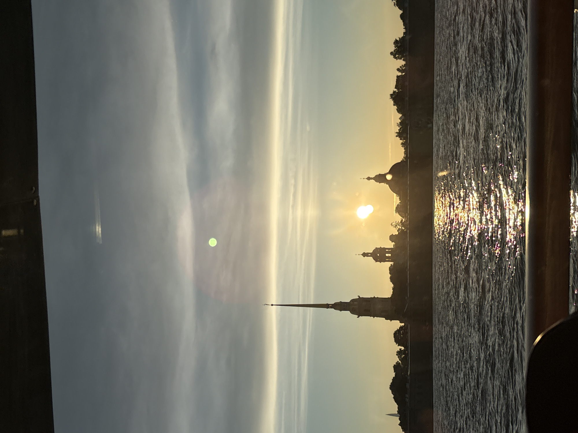 The image depicts a sunset over the Neva River in Saint Petersburg, Russia. In the background, the silhouette of the Peter and Paul Fortress is visible, with its iconic spire and domes. The sun, low on the horizon, casts a golden glow on the water, creating a shimmering reflection. The sky is filled with soft, streaky clouds, transitioning from pale blue to a warm yellow near the sun. The foreground shows the dark, rippling surface of the river, with a glimpse of a dark railing suggesting the viewpoint is from a boat. There are no people visible in the scene. The overall atmosphere is serene and picturesque.