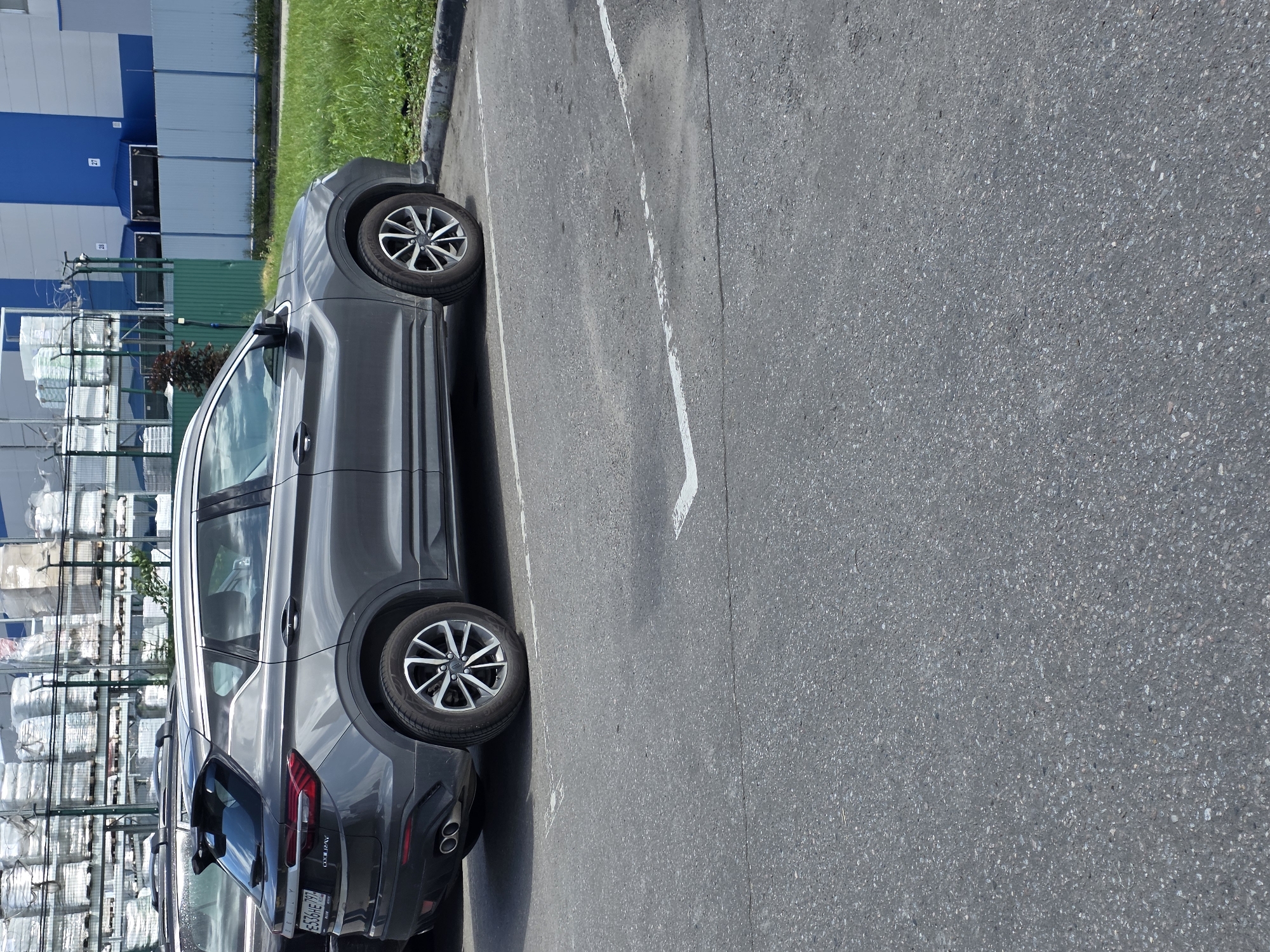 A gray Geely Coolray SUV is parked in an asphalt parking lot. The car is viewed from the rear passenger side. It has chrome dual exhaust pipes and a spoiler on the rear. The license plate reads "E536HE 797". In the background, there is a building with blue and gray siding, stacks of what appear to be building materials on shelves, a green fence, and a patch of grass. The sky is overcast with clouds.