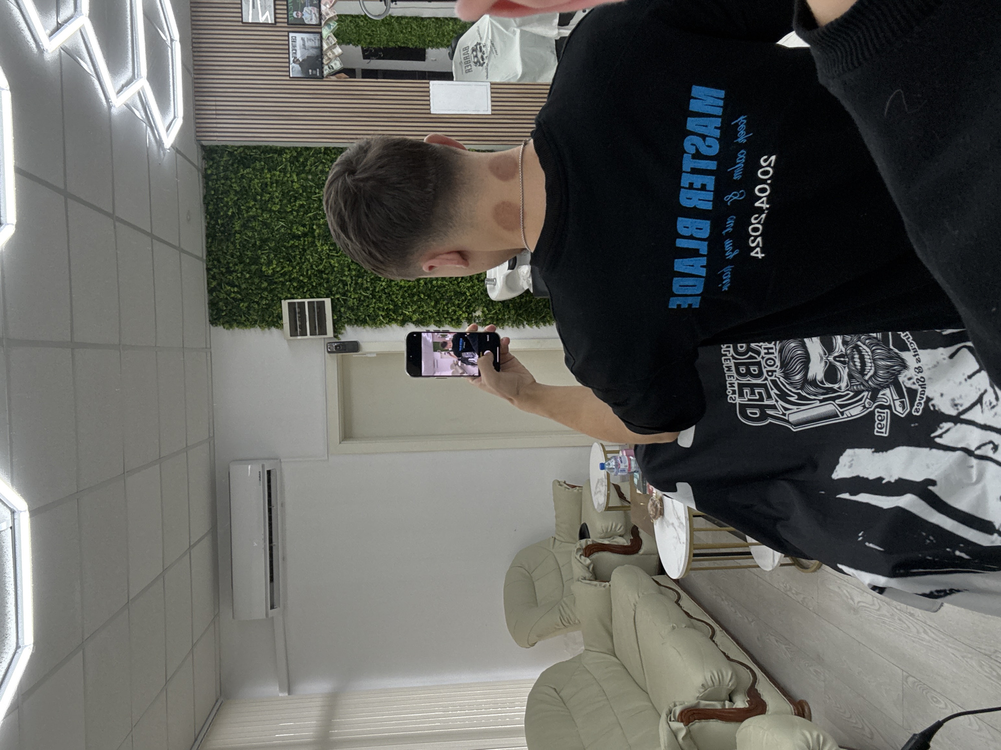 The image captures an indoor scene, likely a barbershop or salon, from behind a male client. The man, who has a fresh fade haircut, is seen from the back. Notably, he has several circular, reddish marks on his neck, consistent with cupping therapy, and wears a silver chain. He is holding an iPhone in his right hand, seemingly taking a photo or video, as the phone screen displays an image of a person with a fresh haircut, possibly himself, alongside another individual (likely the barber).
His black t-shirt bears the mirrored text "MASTER BLADE" in prominent blue capital letters on the back. Below it, other smaller, partially legible mirrored text includes "O.S.O.S.V.O.S.O.S" (suggesting "ASOS"). A black barber's cape or apron, draped over a chair in the foreground, features a stylized skull design with text such as "DEEP" and "1991."
The setting showcases a modern interior design. The background features a decorative green faux-plant wall adjacent to a section with light wooden slats. Above these, framed items, including one with "COURIER" visible, hang on the wall. A white wall-mounted air conditioner is installed near the ceiling. The ceiling itself is fitted with geometric, bright LED lighting. The foreground right shows a waiting area with several light-colored, padded armchairs and small round side tables. The floor is a light-toned wood laminate. The lighting suggests an indoor daytime environment. This establishment is located in Sofia, Bulgaria.