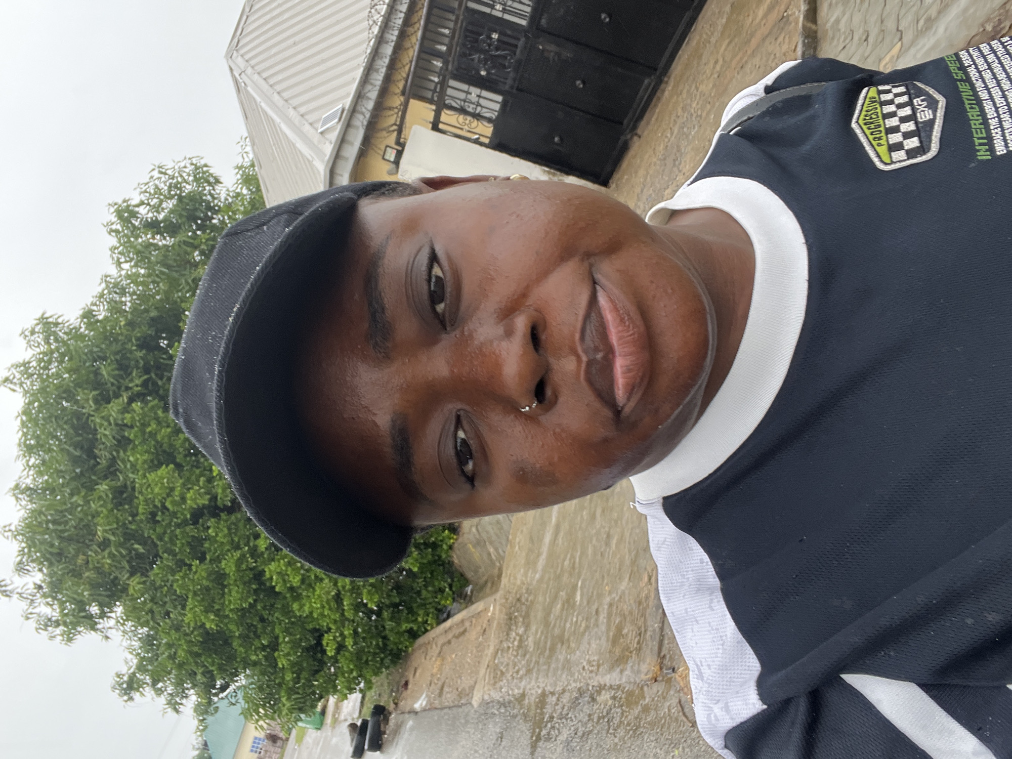 A selfie shows a woman with dark skin, wearing a black baseball cap and a black and white shirt. She has a nose ring and appears to be in an outdoor environment. In the background, there is lush green foliage from a tree, a light-colored roof, and a black metal gate. The ground appears wet, suggesting it may have recently rained, and there are some black objects on the ground in the lower left portion of the image. The overall setting suggests a residential area in Abuja, Nigeria.