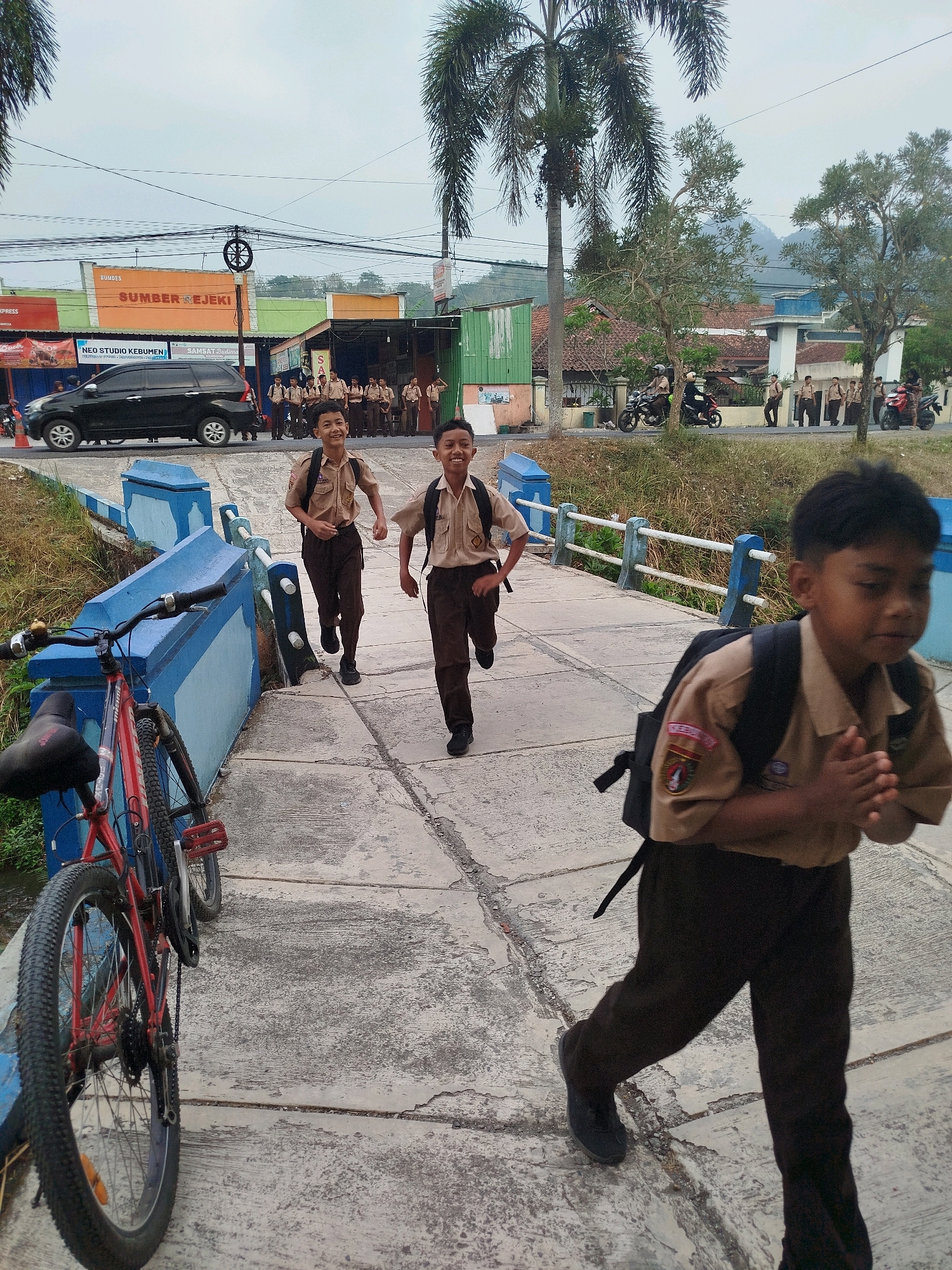 The image captures a scene on a bridge in Kedungwaru, Indonesia, with several young boys in school uniforms running across it. The boy in the foreground, on the right, is partially visible, facing right with his hands clasped. He wears a tan uniform shirt with a patch on the sleeve and dark brown pants, and carries a black backpack. Two other boys in similar uniforms are running ahead of him on the bridge. The boy on the left is further back, while the boy in the middle is closer to the viewer. They are all in motion, with their legs blurred from running.
The bridge has a light-colored concrete surface with visible cracks and is bordered by blue and white railings. To the left of the bridge, a red bicycle is parked, leaning against the railing. The background shows a street with a black car, several motorcycles, and a line of children standing on the sidewalk, also in school uniforms. Buildings with signage, including "SUMBER REJEKI" and "NEO STUDIO KEBUMEN," are visible behind the children. Palm trees and other vegetation are present, and a mountainous landscape can be seen in the distance. The sky is overcast, suggesting it might be daytime with cloudy weather.