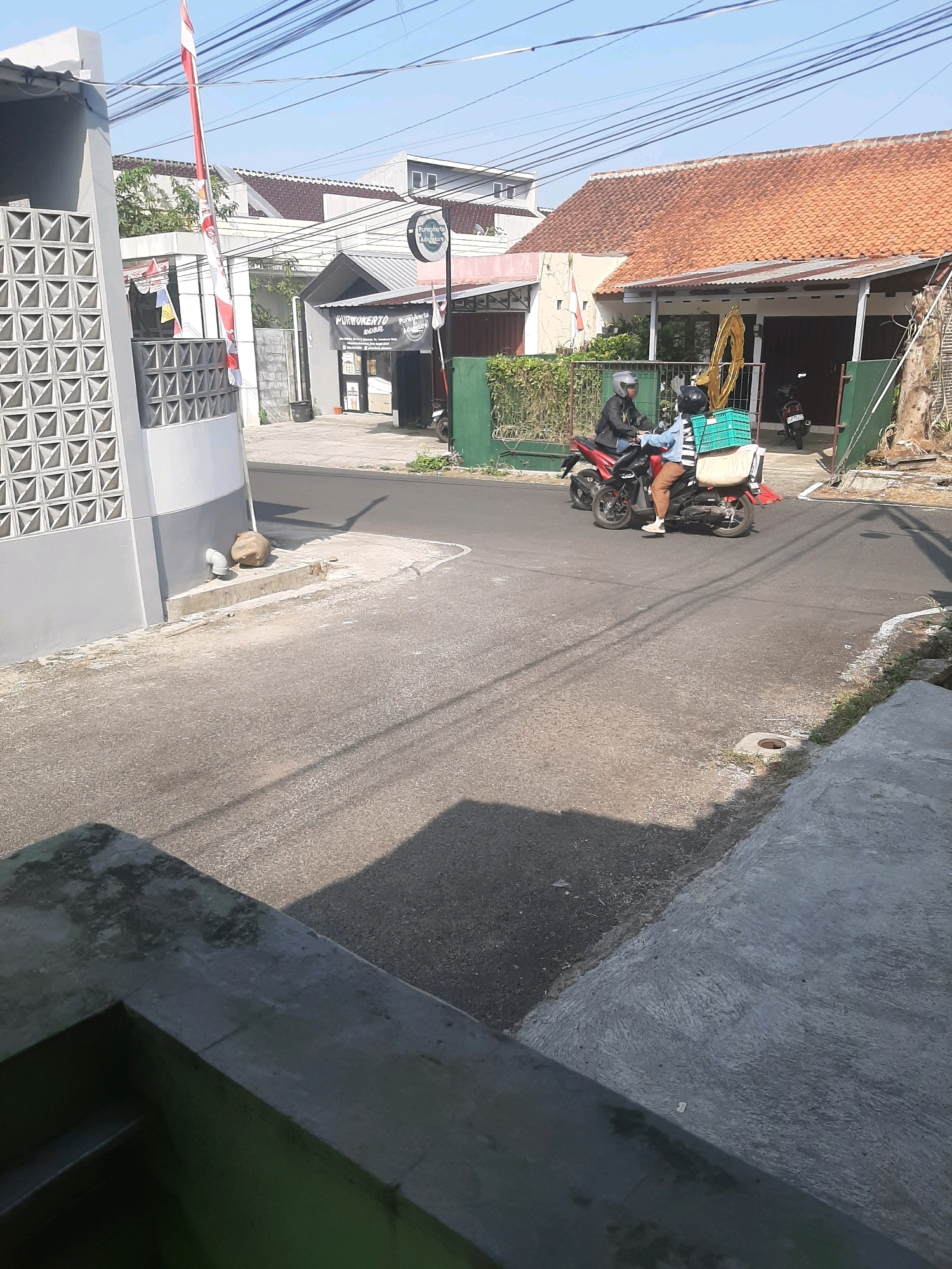 In Sumampir, Indonesia, on a sunny day, a street scene unfolds with a light-colored building on the left featuring a decorative grey wall. Adjacent to it, a red and white Indonesian flag flies from a tall pole. Further down the street, a building with a terracotta tile roof displays a sign for "PURWOKERTO ADVENTURE." Two individuals on a red scooter are in motion. The driver, wearing a black helmet and jacket, is visible, while the passenger behind, also helmeted, wears a blue jacket and brown pants. They are carrying a large cream-colored bag and a green crate on the back of the scooter. Another scooter is parked in front of the building on the right. The road is asphalt, with a concrete sidewalk and some scattered debris. The surrounding environment suggests a residential or commercial area with overhead power lines.