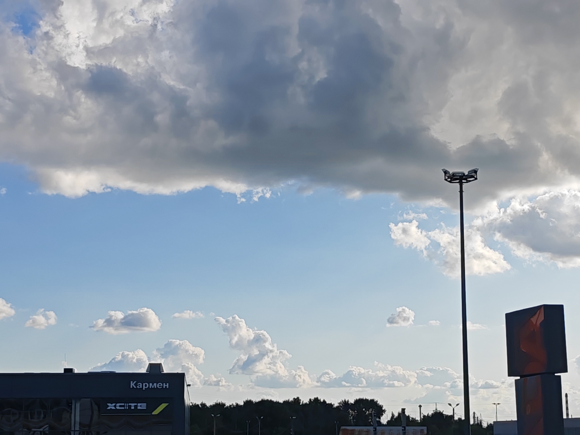 The image depicts a vast sky with a mix of dark and light clouds, typical of an overcast day. In the foreground, on the left side, is the corner of a dark building with the word "Кармен" (Carmen) written in white Cyrillic letters above a logo that reads "XCITЕ". To the right of the building, a tall lamppost with four spotlights stands prominently. Further to the right, a large dark cube-shaped sign is visible. In the background, a line of trees forms a dark silhouette against the sky. The scene appears to be an outdoor urban or industrial area.