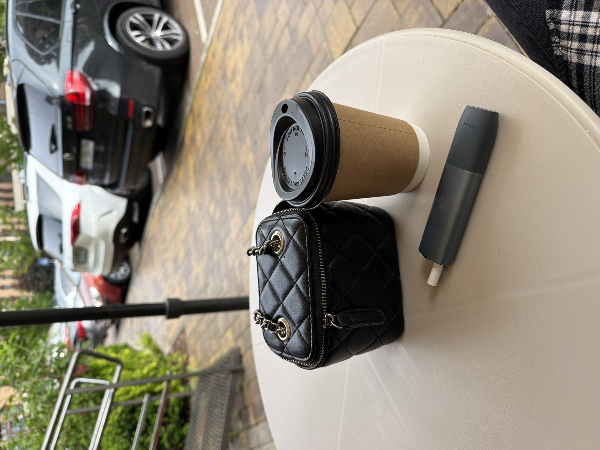 The image provides an overhead view of personal items arranged on a light-colored, round outdoor table, set against a blurred background of a street scene and greenery.
**Subjects:** On the table, from left to right, are a brown paper coffee cup with a black plastic lid, a black quilted mini bag with gold-toned chain details and a zipper closure, and a dark grey IQOS device. A small portion of a person's leg or clothing, featuring a black and white plaid pattern, is visible on the far right edge of the table. The blurred background shows several cars, including a black SUV, a white car, and a red car. Green foliage, a black metal railing, and a patterned paved ground are also discernible.
**Activity or event taking place:** The arrangement of these items suggests a casual break or a moment of relaxation, likely at an outdoor cafe or a balcony overlooking a street. No direct human activity is shown, but the presence of the items implies a personal pause.
**Notable details:** The scene appears to be set during the daytime. The lighting suggests it might be an overcast day or late afternoon. The location is Sofiivska Borshchahivka, Ukraine, as indicated by the context and the Ukrainian text on the coffee cup.
**Visible text:**
* On the black lid of the coffee cup, in white letters, "CAUTION HOT" (upside down), "34 90", "06", and "ОБЕРЕЖНО ГОРЯЧЕ" (Ukrainian for "CAREFUL HOT").
* The dark grey device has the "IQOS" logo embossed on its upper section.
* A license plate on the black SUV in the background is partially visible, appearing to be a Ukrainian plate, possibly starting with "AI".