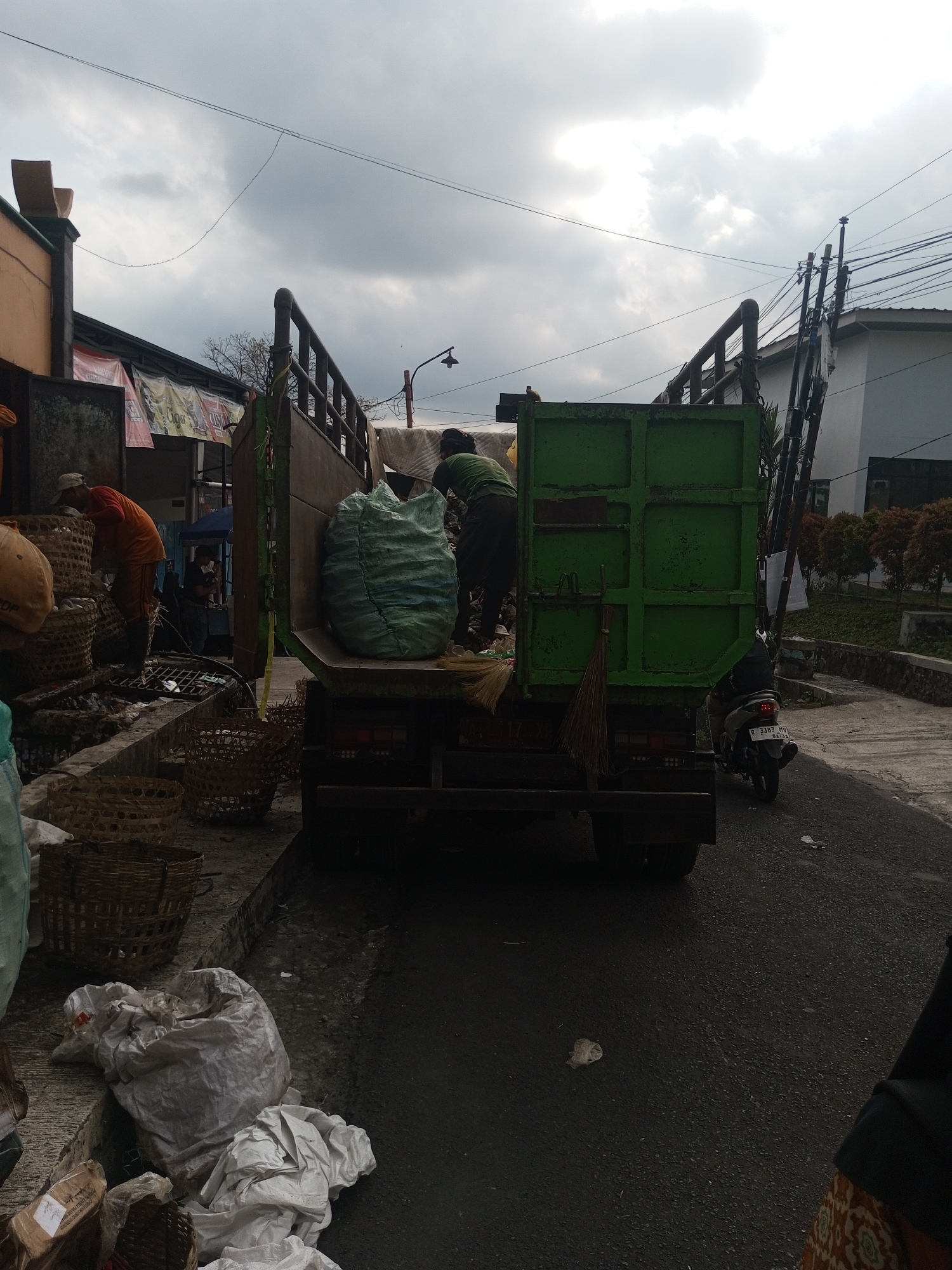 In Krasak, Indonesia, a garbage truck is being loaded by two men. One man, wearing an orange shirt and a cap, is standing and appears to be sorting through refuse. The other man, wearing a green shirt, is bent over and actively placing a large, light blue, woven bag of trash into the back of the green truck. The truck bed is open, revealing a substantial amount of garbage. In the foreground, several woven baskets and large white plastic bags filled with waste are piled on the ground. A broom leans against the side of the truck. A motorcycle is parked on the right side of the road, facing away from the truck. The scene takes place on a paved road under a cloudy sky, with buildings and trees visible in the background.