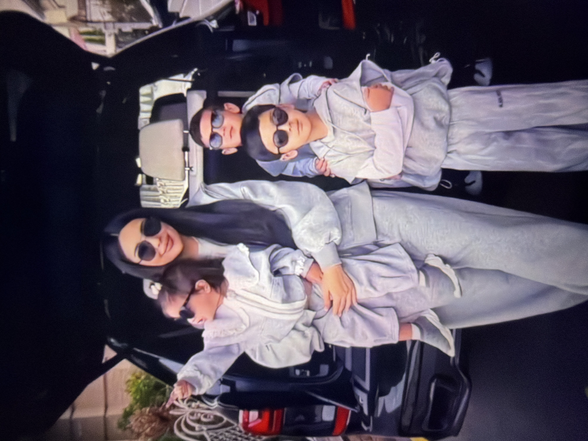 In Beșalma, Moldova, a family of four poses in front of a black SUV with its tailgate open. A woman, appearing to be the mother, stands center, holding a baby on her left arm. She has long, dark hair and wears sunglasses and a light grey tracksuit. The baby, also wearing sunglasses and a light grey outfit, points to the left. To the right of the woman, two boys stand, also in light grey tracksuits and sunglasses. The older boy is behind the younger boy, with his arm around the younger boy's shoulders. The SUV's interior is visible, with light-colored headrests. The background suggests an outdoor setting, possibly a residential area, with some greenery and architectural elements visible.