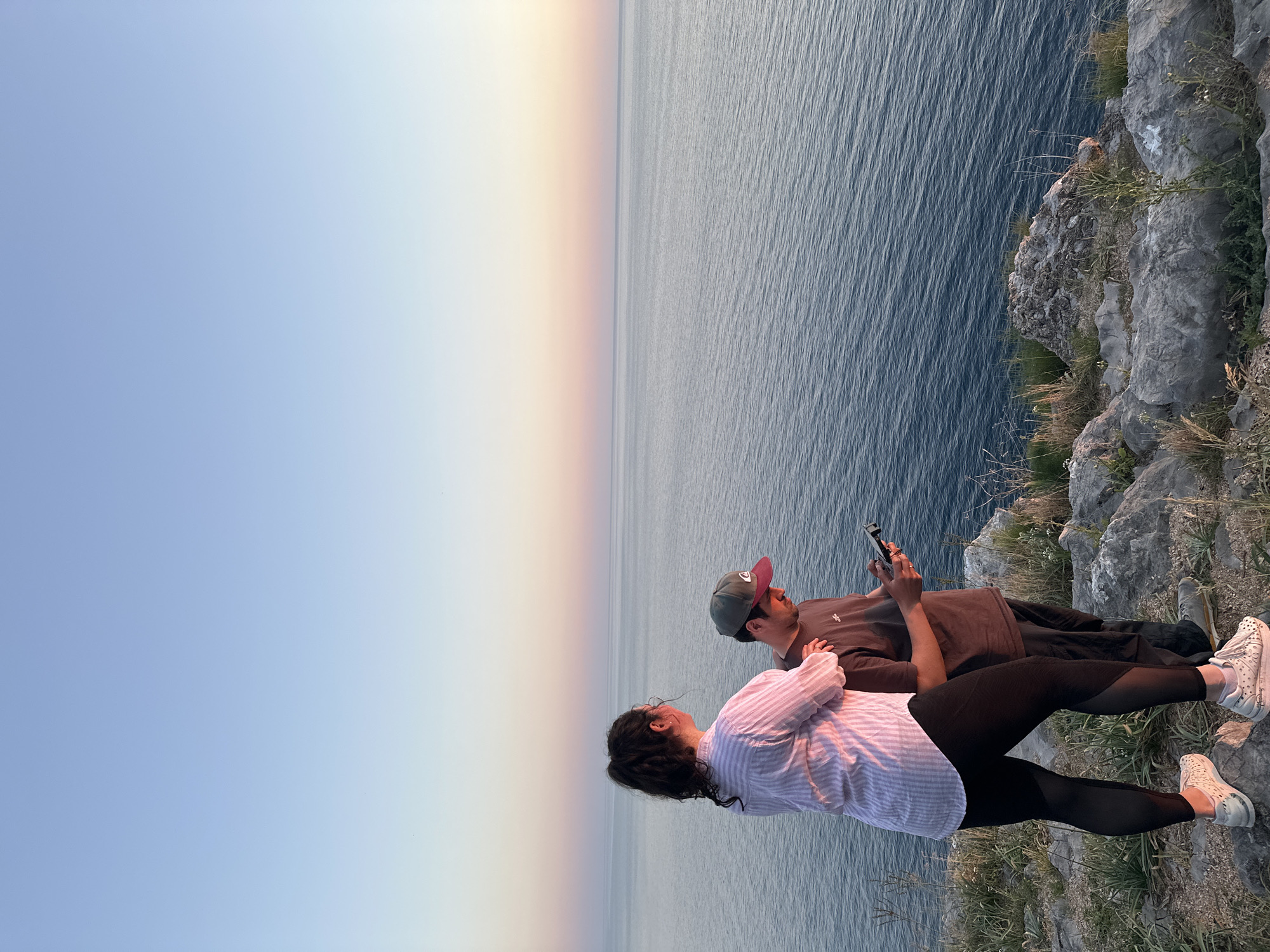 Two people are standing on a rocky cliff overlooking the sea at sunset, in Petrovac centar, Montenegro. One person is wearing a striped shirt and black leggings. The other person is wearing a dark shirt and a baseball cap and appears to be holding a device. The cliff is covered in grass and the sky is light blue blending into a sunset hue.