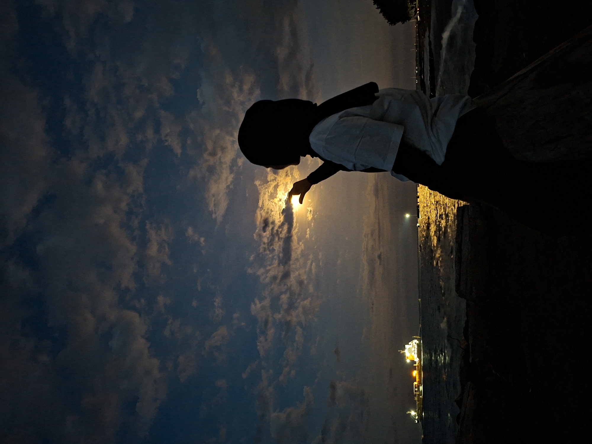 A low-angle shot in Lhokseumawe, Indonesia shows a silhouetted person wearing a head covering standing on a dark, rocky surface near the water. The person extends a finger towards a bright, slightly obscured sun or moon in the sky. The sky is filled with clouds, transitioning from a dark blue on the left to lighter shades near the horizon, with golden highlights around the celestial object. Across the water, lights indicate the presence of buildings and possibly a ship or other structures, creating reflections on the water surface.