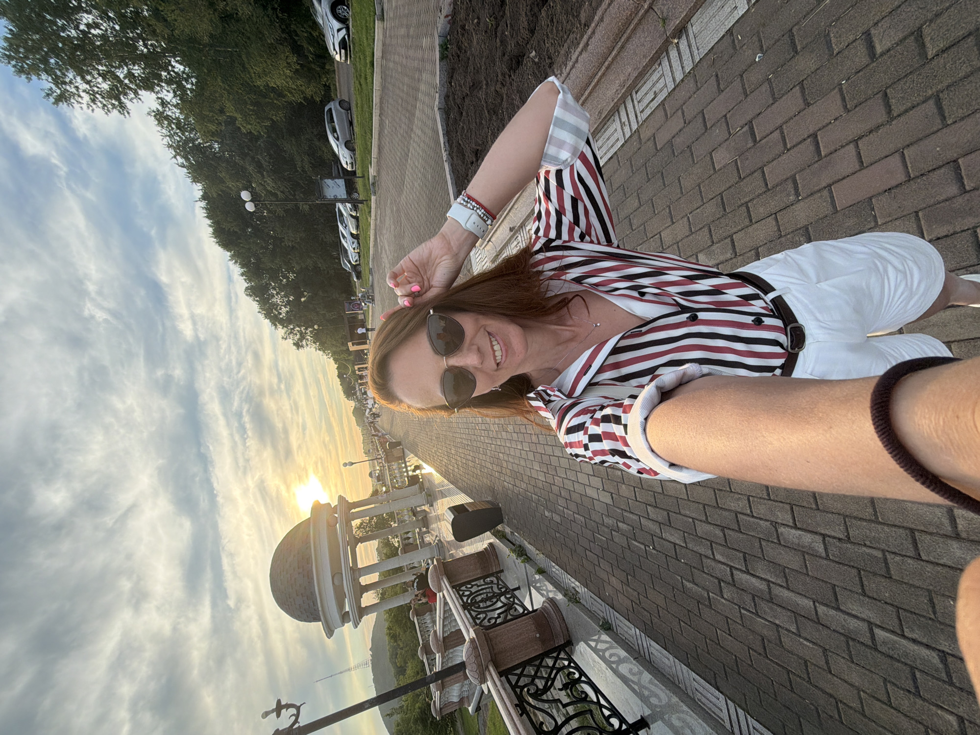 The image shows a woman taking a selfie outdoors in Birobidzhan, Russia, likely during sunset. The woman is wearing a striped shirt and white shorts. She is standing on a paved walkway lined with decorative railings and a gazebo in the background. A row of cars is parked along the side of the walkway, and trees and buildings are visible in the distance. The sky is cloudy with the sun setting.