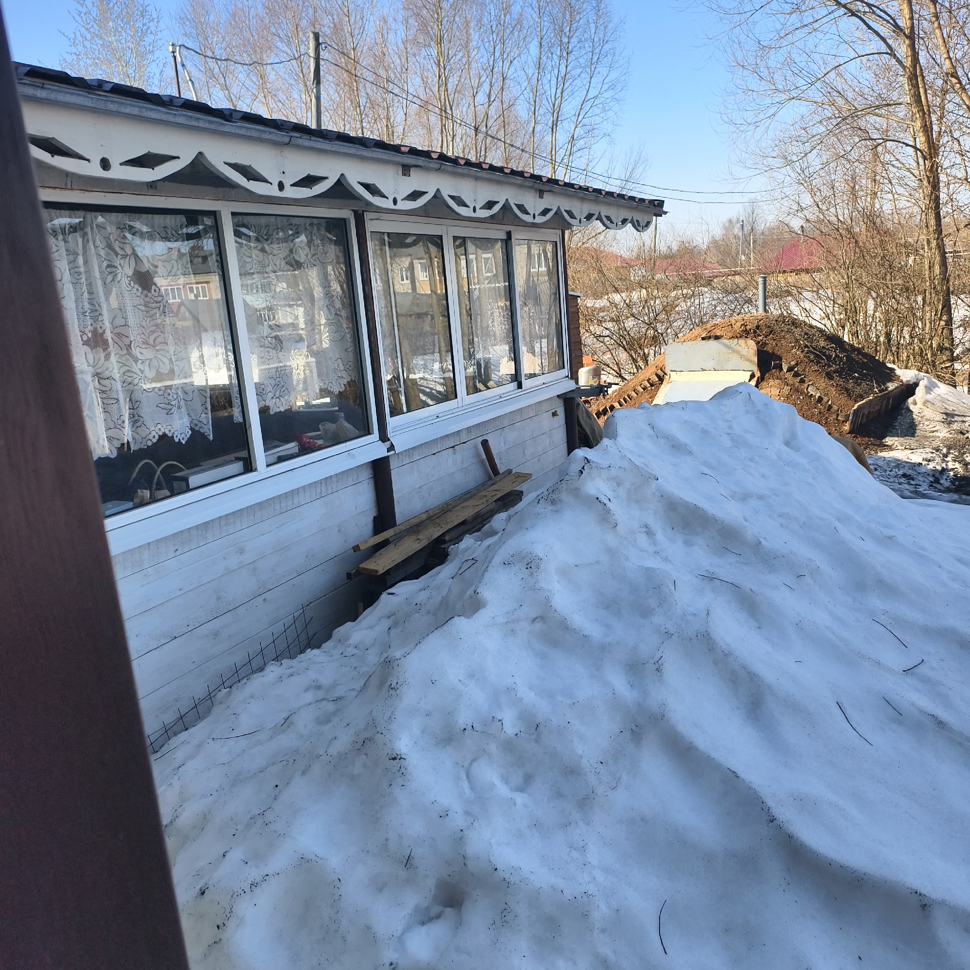 The image shows a white, single-story building with large windows covered by lace curtains. A pile of snow is situated in front of the building, with wooden planks resting against the wall and partially buried in the snow. A wire fence is visible along the base of the snow pile. In the background, there is a pile of dirt and vegetation, with bare trees reaching toward a clear blue sky. A red-roofed building is also visible in the distance. The image appears to be taken during the day in Фоки, Russia. No people are visible.
