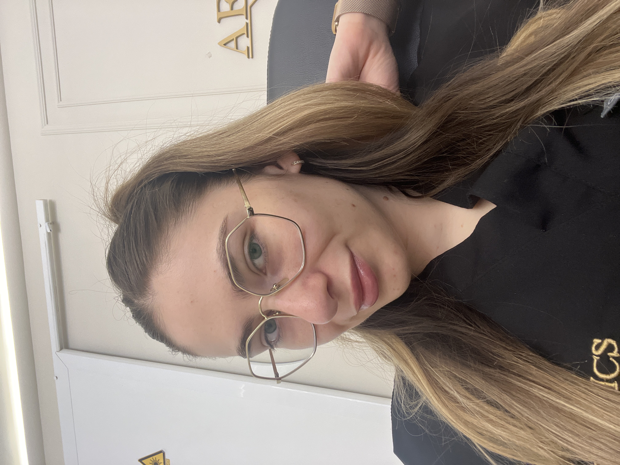 A woman with long, light brown hair pulled into a ponytail is looking at the camera. She is wearing large, hexagonal, gold-rimmed glasses and a black shirt with gold lettering that reads "ICS". She has a small hoop earring in her left ear and is wearing a rose gold watch on her right wrist. Her right hand is resting on the back of her head. The background appears to be an indoor setting with white walls. A yellow warning symbol is visible on the left side of the image. Gold lettering, partially obscured, is visible on the wall to the right.