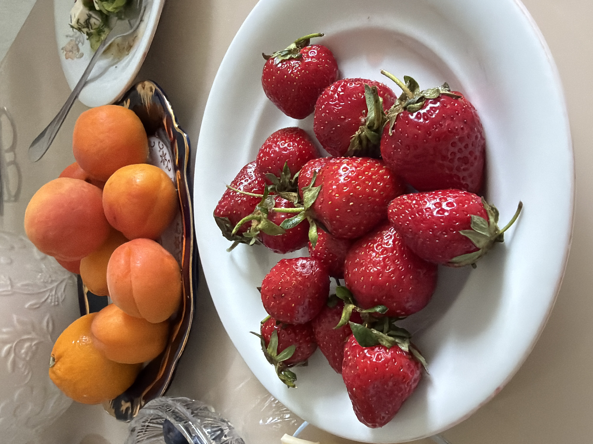 A table is set with several plates of fruit. One plate contains apricots in a decorative gold and blue dish. Another plate is filled with fresh strawberries. A third plate contains salad and a fork. The location is not discernible from the image.
