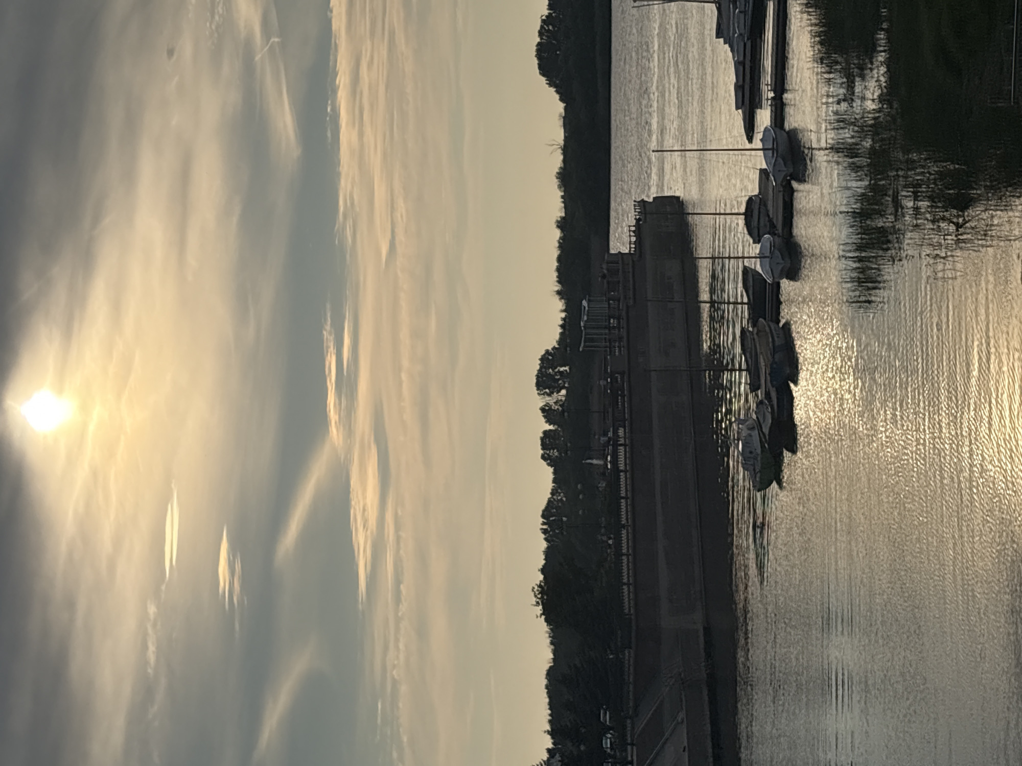 The image depicts a waterfront scene in Astrakhan, Russia, during daylight, likely late afternoon or early evening, given the soft, golden sunlight illuminating the sky and water. Dominating the upper portion of the frame is a hazy sky with scattered clouds, through which the sun is visible as a bright, diffused orb. The foreground features a body of water, possibly the Volga River, with gentle ripples reflecting the sky's light.
Along the left side of the water, a long, flat-bottomed barge or pier structure is moored. Several small sailboats are docked alongside wooden piers extending from the shore or barge. These sailboats are mostly white or light-colored, with masts and rigging visible. Some are tied together, while others appear to be moored independently. On the right side of the frame, more boats are visible, some resting on a floating dock.
Behind the waterfront, a dense line of trees forms a dark silhouette against the lighter sky. In the distance, faint lights and structures are barely discernible through the foliage, suggesting an urban or park area. A portion of a concrete embankment or river wall is visible in the lower left quadrant. The overall atmosphere is serene, with a sense of tranquility conveyed by the calm water and the soft lighting. There are no people clearly visible in the frame.