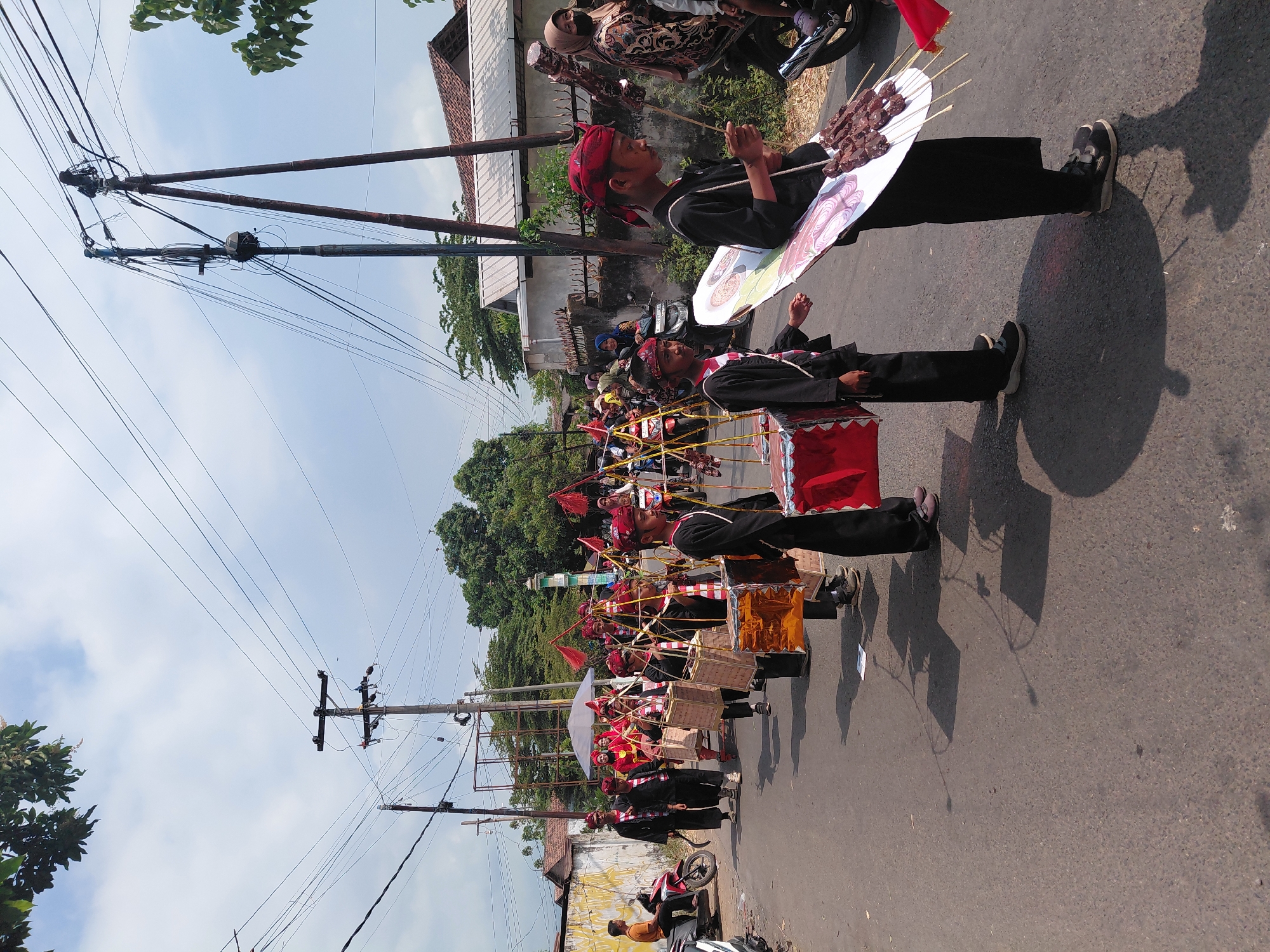 In Bimo, Indonesia, a procession is taking place on a paved road under a partly cloudy sky. The group consists of young people, mostly boys, dressed in traditional black attire with red headbands and sashes. They are carrying various items, including square wooden boxes adorned with red and gold paper, some of which appear to be decorated with intricate patterns. One boy in the foreground is holding a large, flat, decorated object that resembles a fan or a shield, and is skewered with what looks like meat on sticks. Another boy is carrying a similar object, also with meat skewers.
The procession is moving from left to right, with spectators visible in the background. Some individuals are watching from the side of the road, while others are on motorcycles. There are utility poles with numerous wires running overhead, suggesting an urban or semi-urban setting. Buildings with tiled roofs and walls are visible behind the procession. The overall atmosphere suggests a cultural event or a parade.